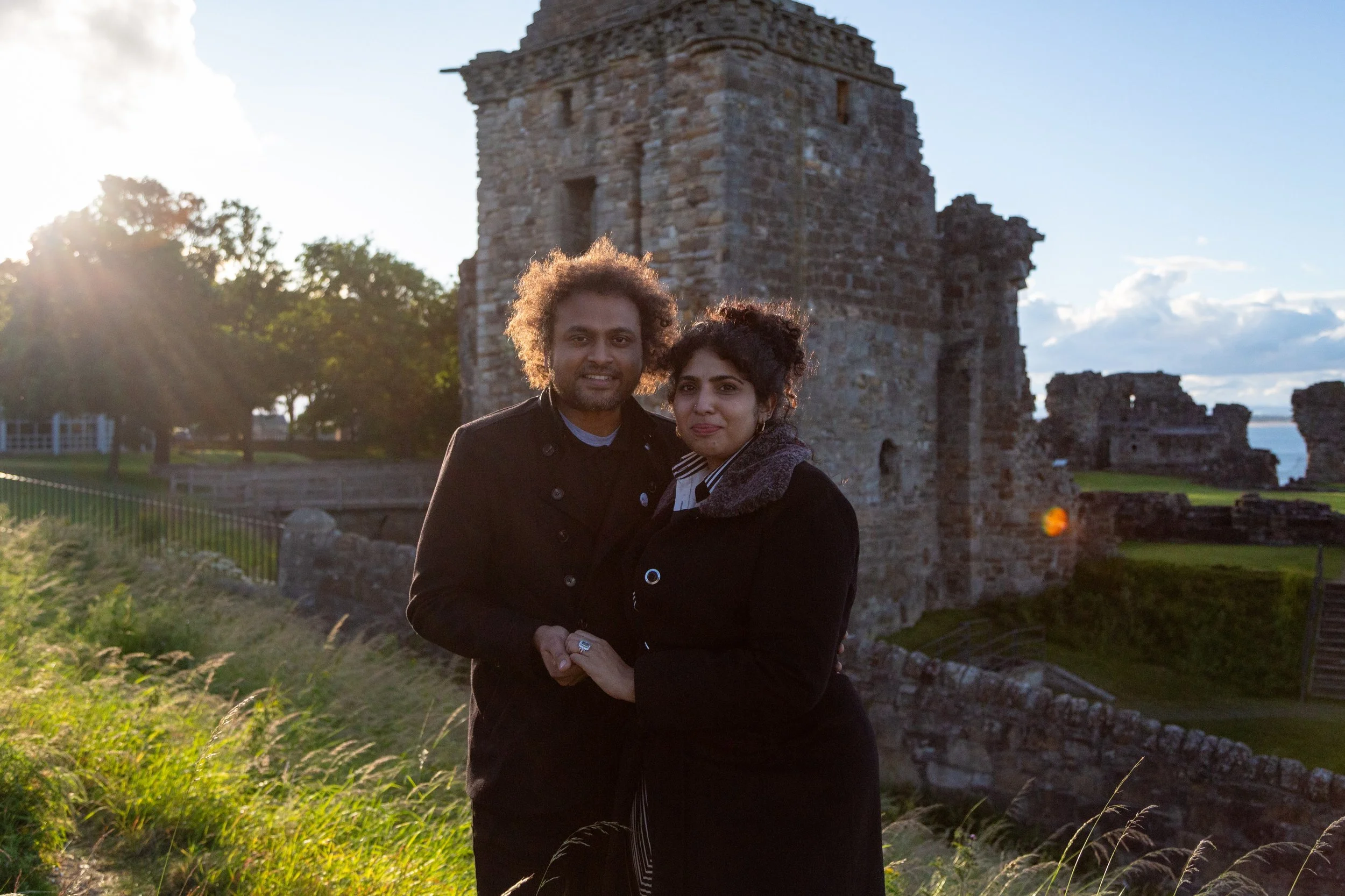 A couple standing close together outdoors during sunset, with an old stone building and trees in the background.
