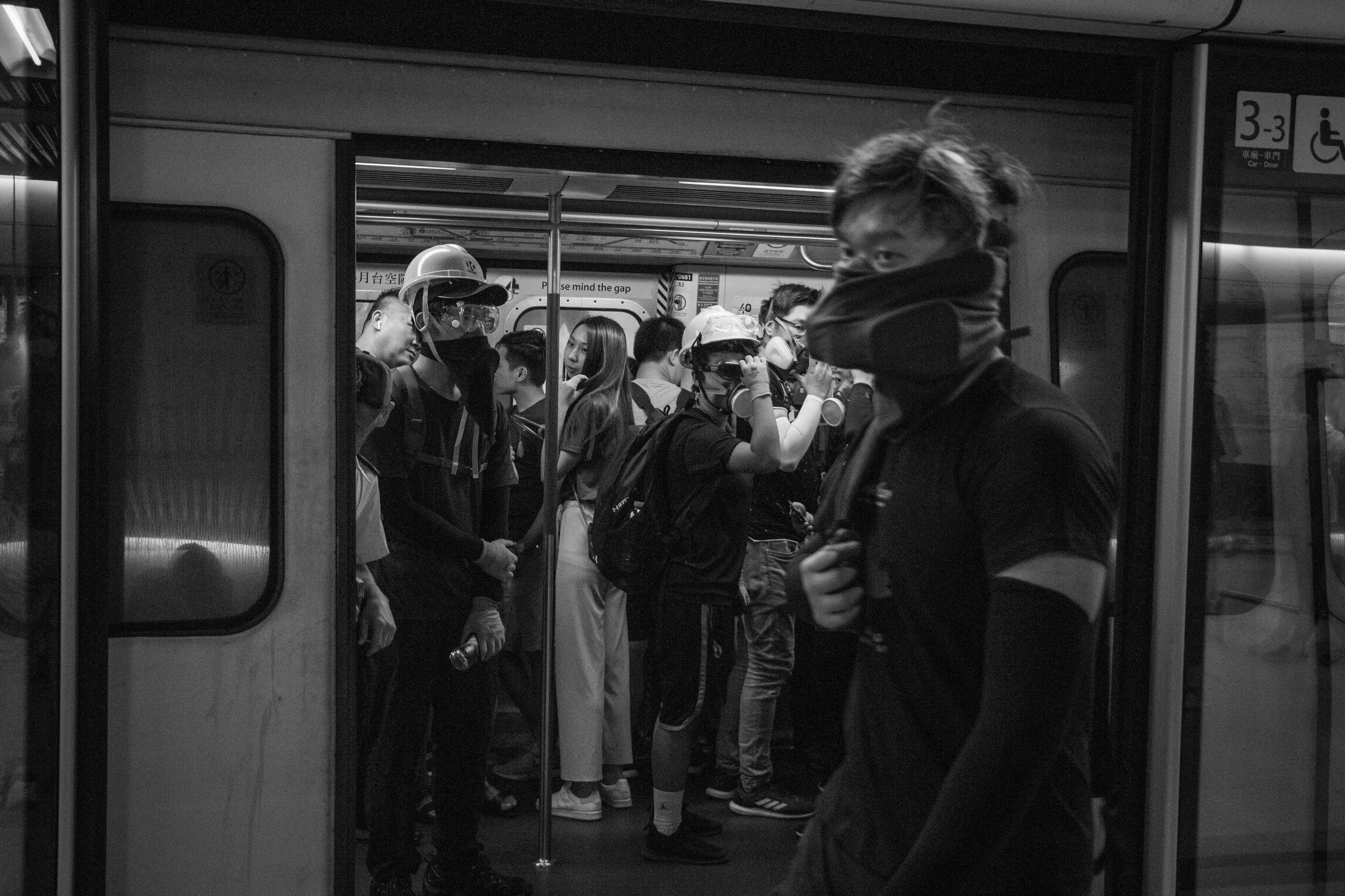 People standing in line inside a subway train station, some wearing masks and helmets, waiting for their train.