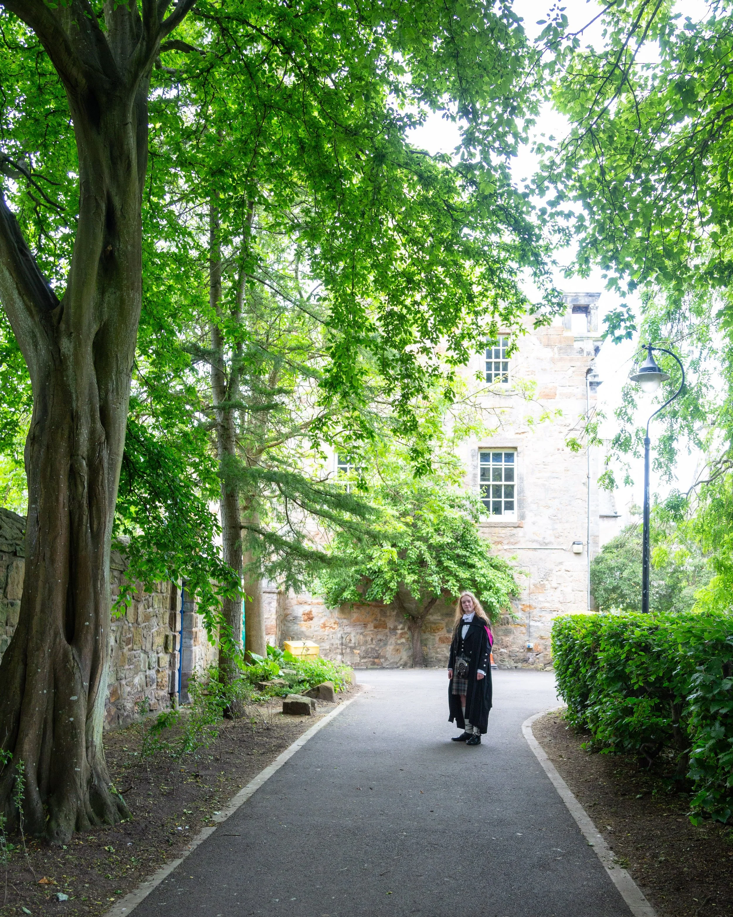 A person in a black graduation gown and cap standing on a winding pathway in a park-like setting, surrounded by lush green trees and bushes, with a stone building and streetlamp visible in the background.