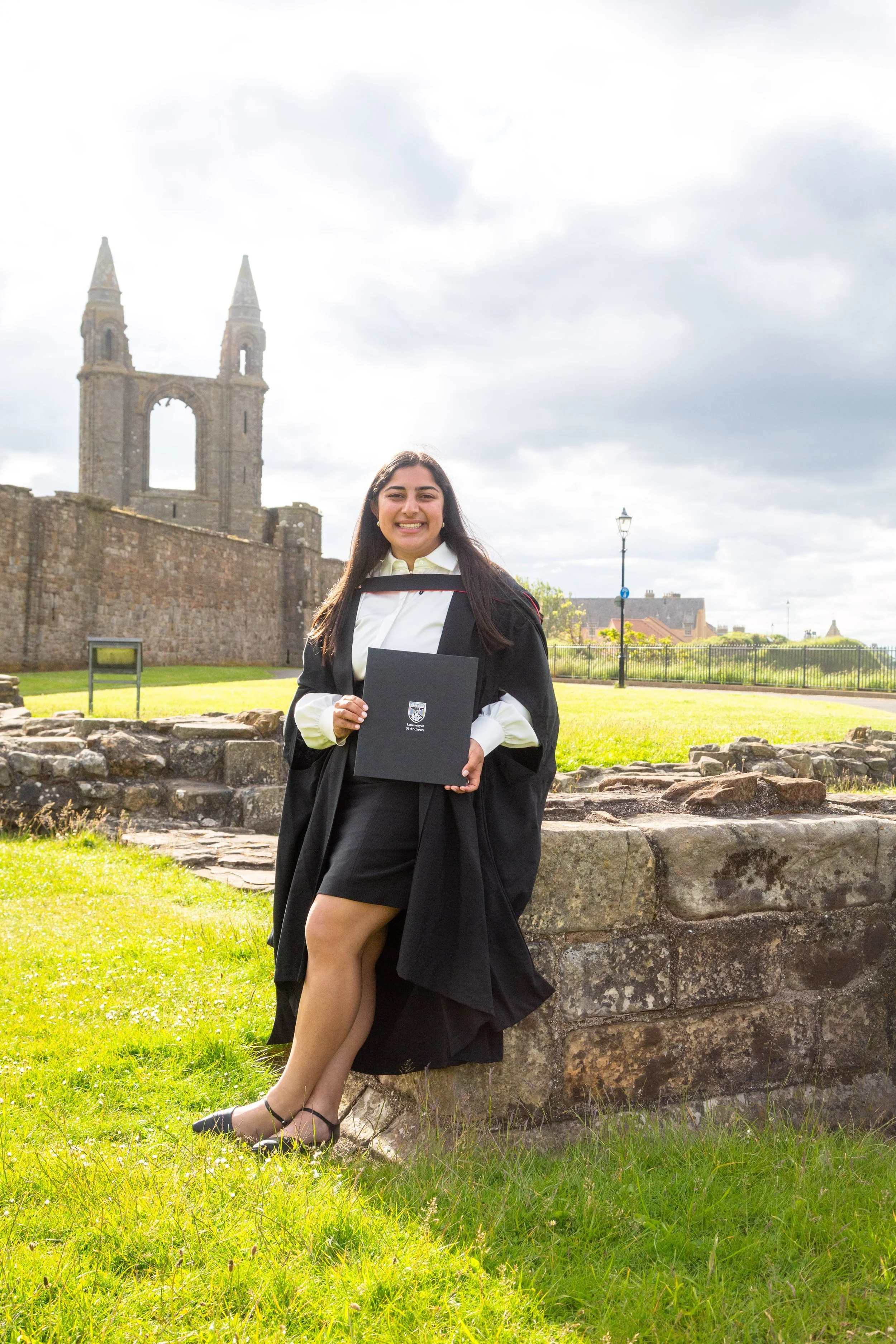 A young woman in a graduation cap and gown standing outdoors on a stone wall, holding a diploma, with a historic stone building and cloudy sky in the background.