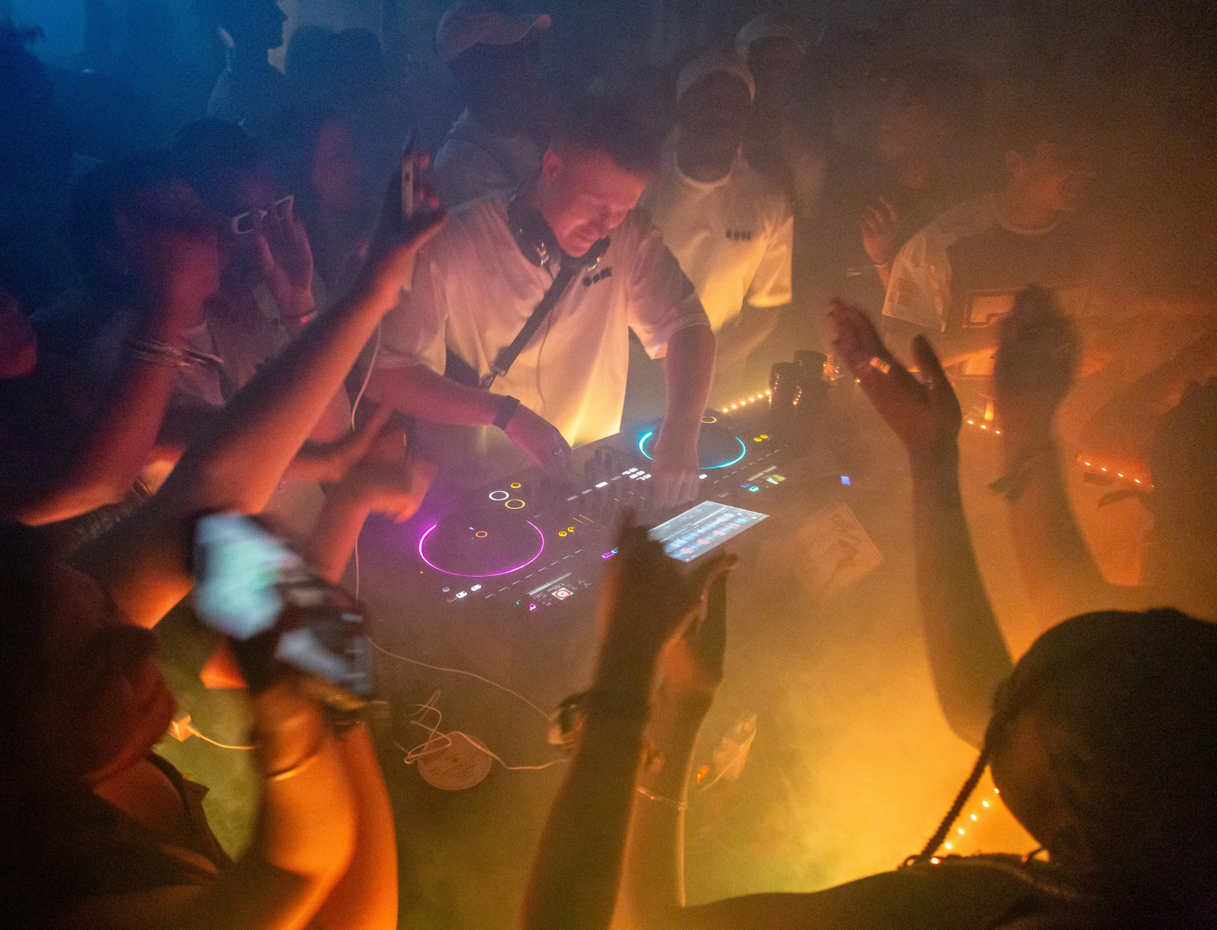 A DJ wearing a beige shirt with a logo, using DJ equipment with colorful lights, surrounded by a crowd in a smoky, dimly lit environment. People are raising their hands and engaging with the music.