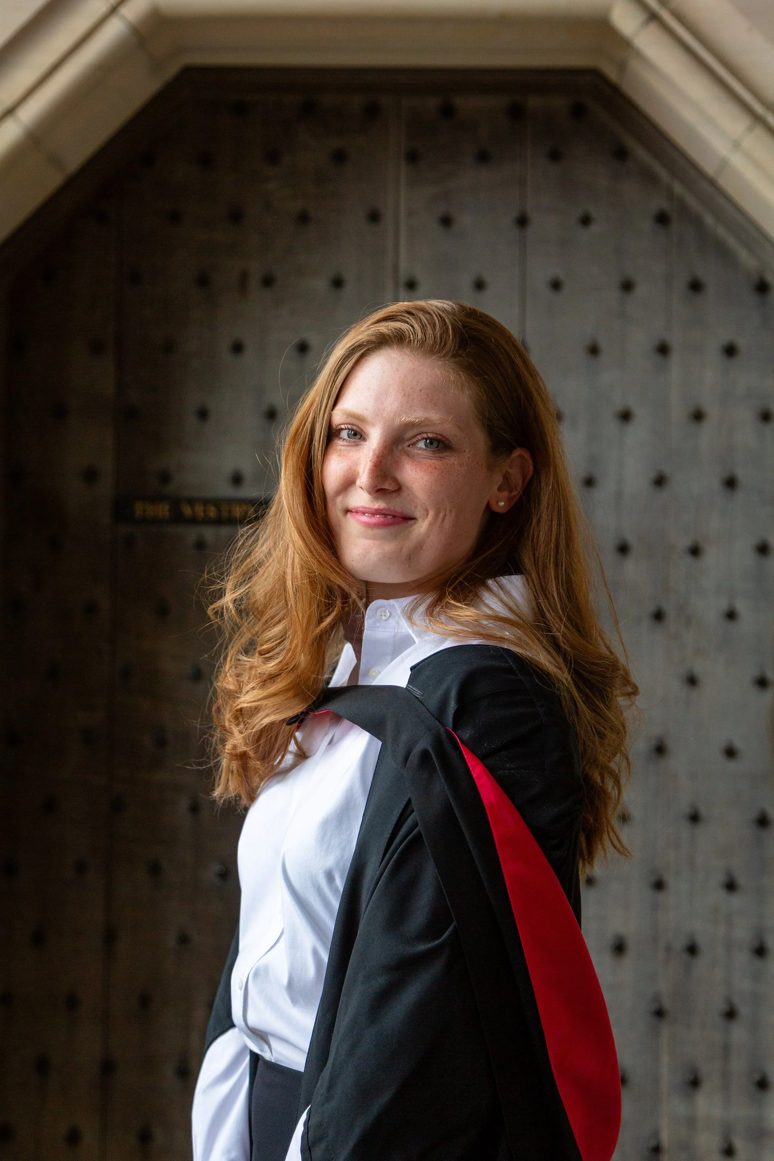 A young woman with long red hair and freckles in a white shirt and black graduation gown posing in front of a dark, textured wall.