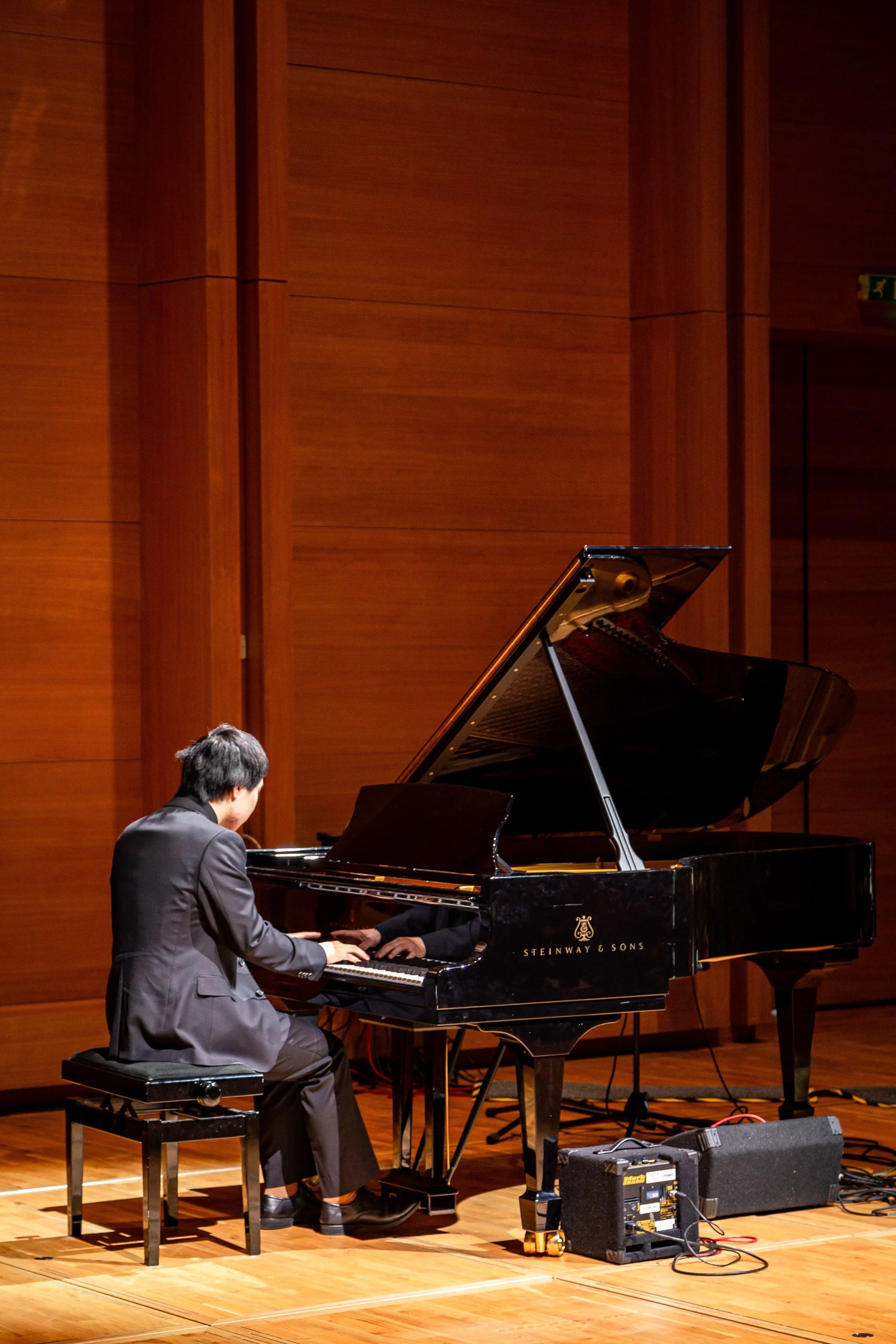 A man in a black suit playing a grand piano on a stage with wooden walls.