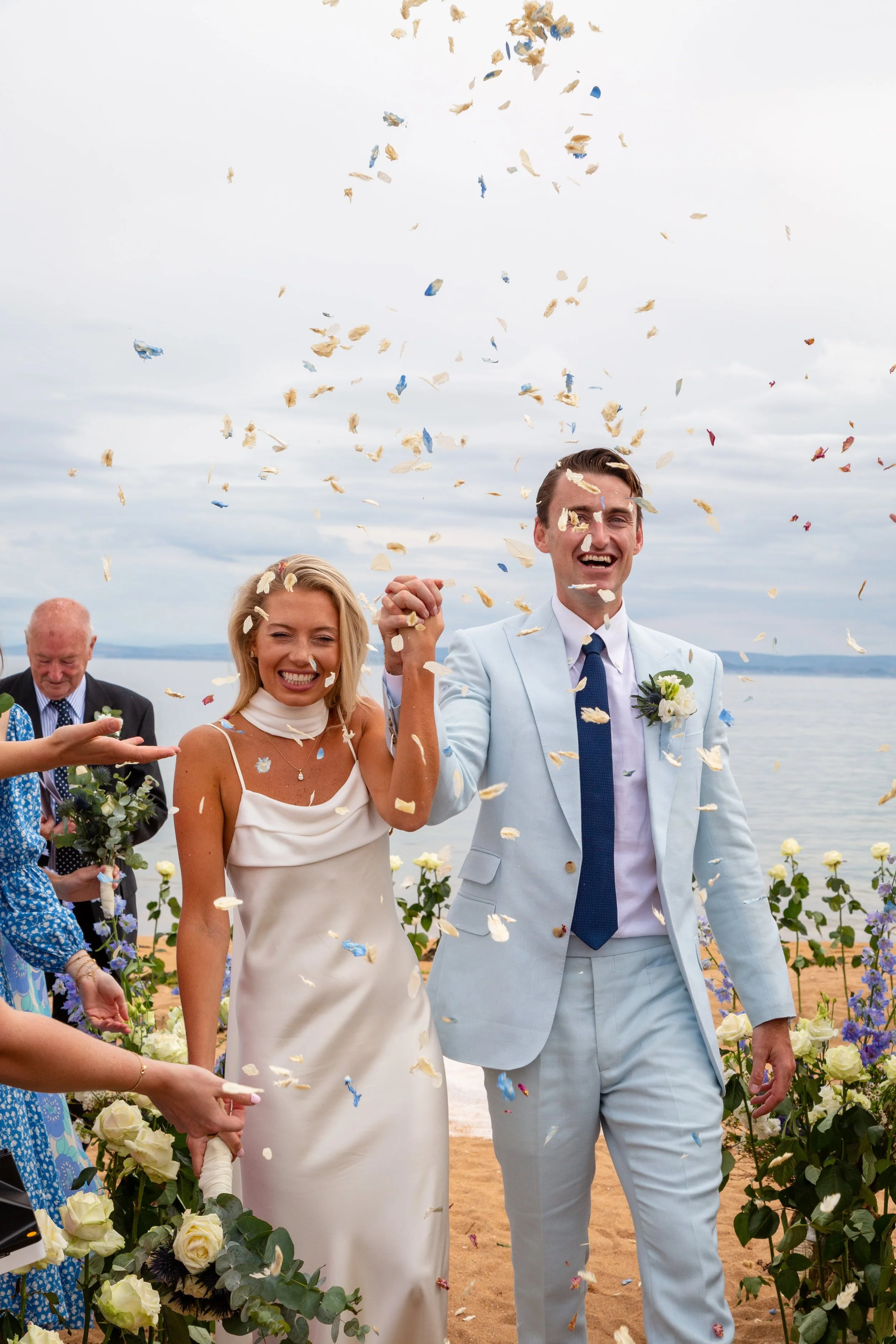 A smiling bride and groom holding hands during their outdoor wedding ceremony on a beach, with confetti falling around them and ocean in the background.