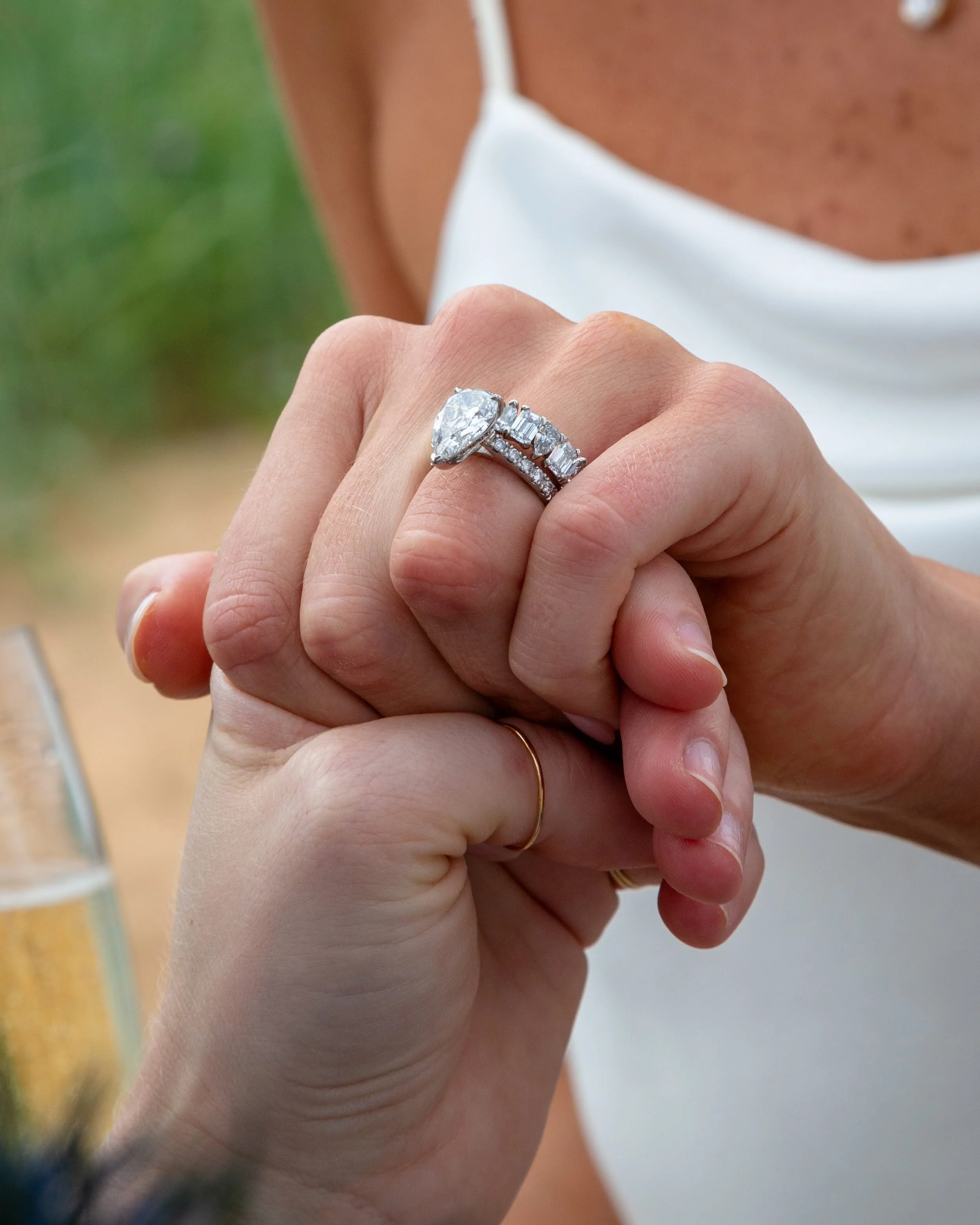 Close-up of a hand showing two rings, one large diamond ring and a band with smaller diamonds, on the finger of a person wearing a white dress with thin straps.