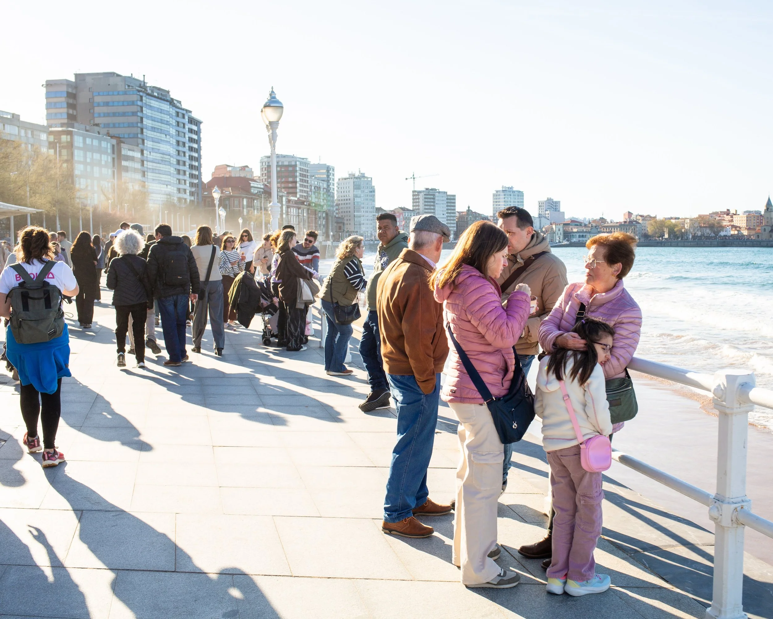 People standing along a city riverfront promenade, with high-rise buildings and city skyline in the background during daylight.