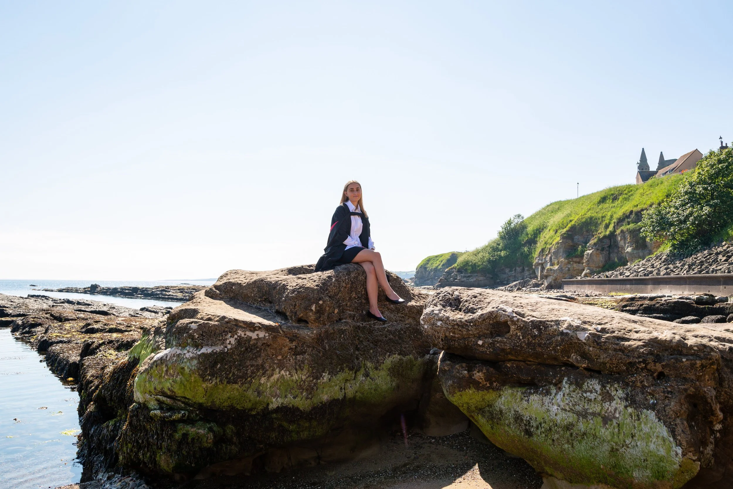 A woman in a black blazer and skirt, white blouse, and black shoes sitting on a large rock near the shoreline with a castle or large building on a green hill in the background.