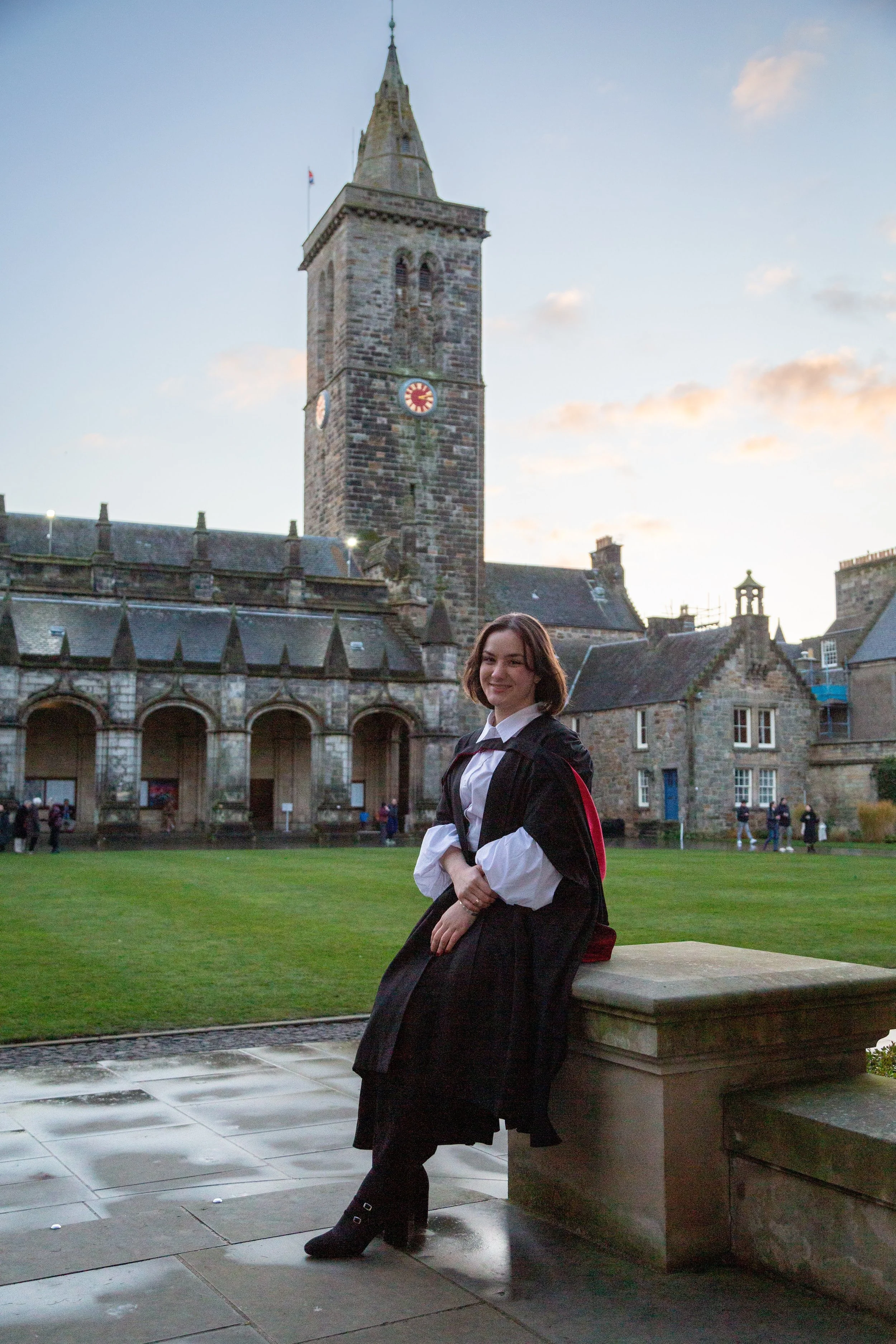 A young woman in a graduation gown standing outdoors on a stone ledge with her arms crossed, smiling, with historic stone buildings and a clock tower in the background during the evening. The sky is partly cloudy.