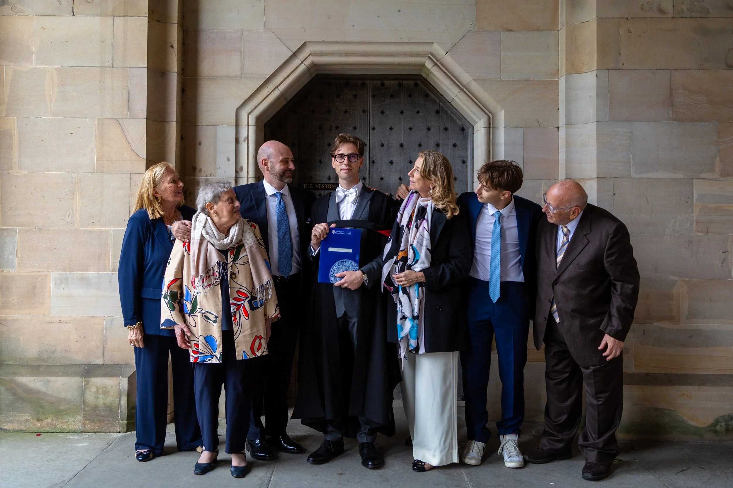 A group of seven people gathered inside a building with stone walls, with one person in graduation cap and gown holding a diploma, flanked by other individuals in formal attire.