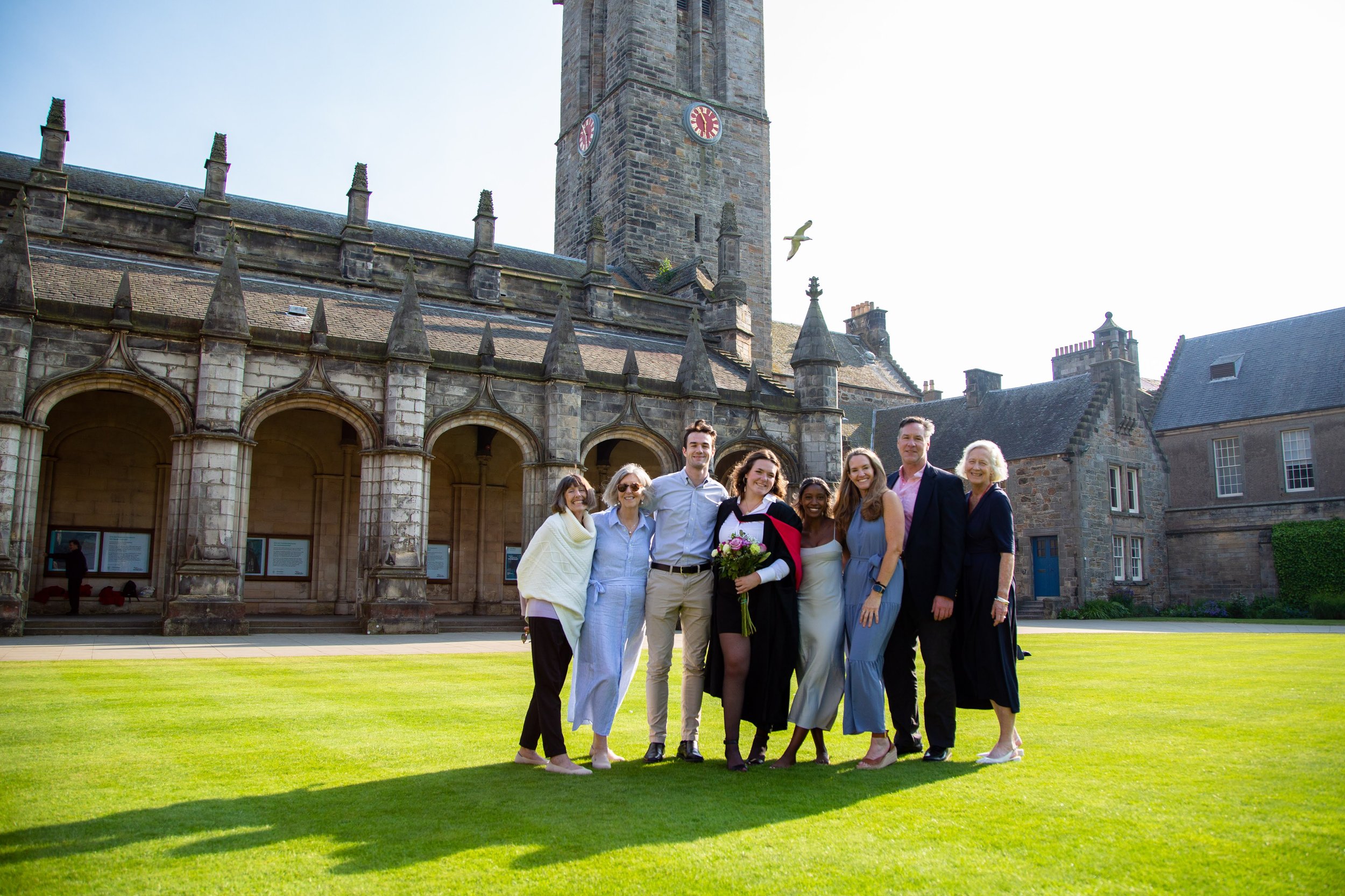 A group of diverse people standing together outdoors in front of an old stone building with a clock tower, celebrating a special occasion with a woman in a graduation cap holding a bouquet of flowers.