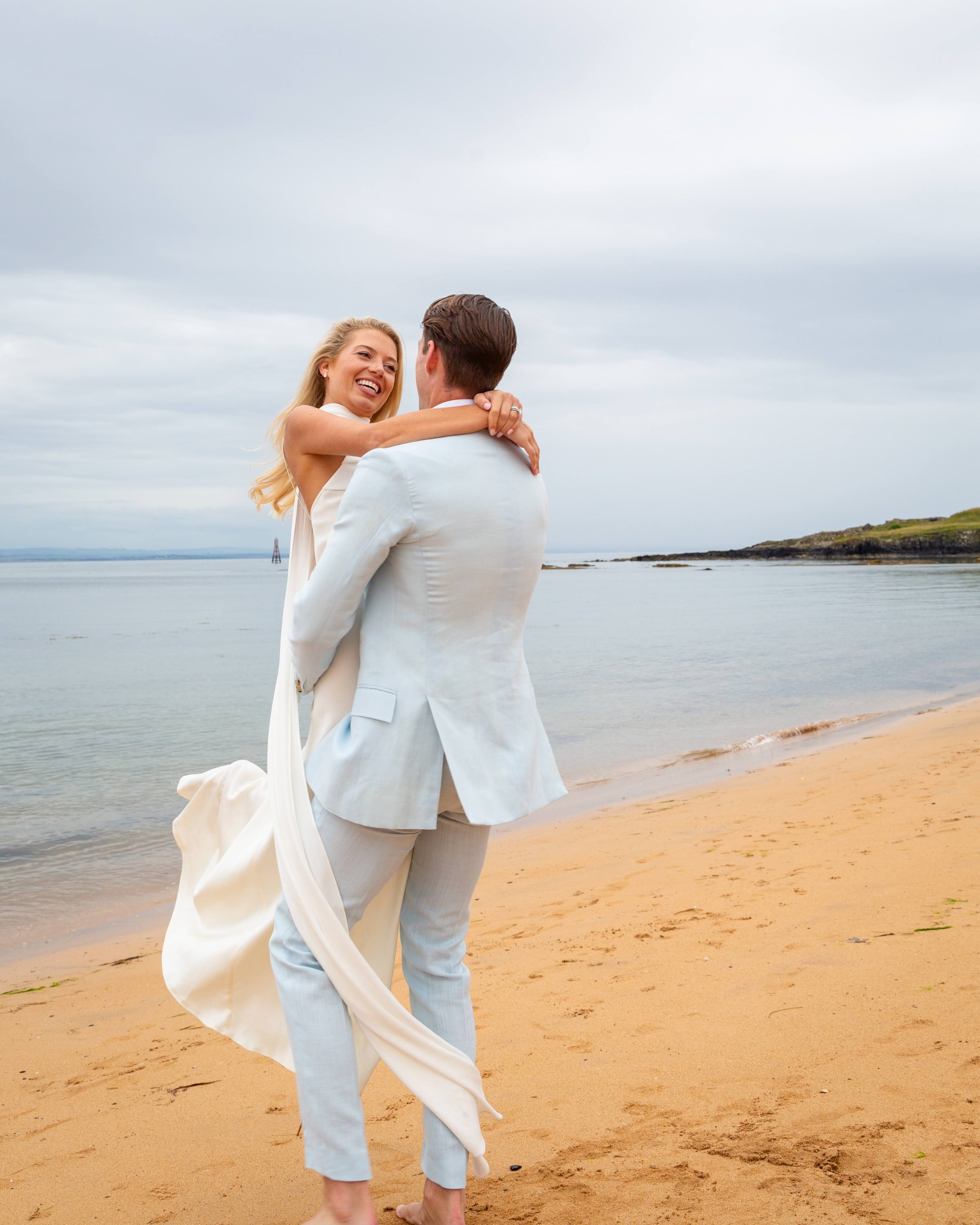 A couple dressed in white is on a beach, with the woman smiling and hugging the man. The background includes the ocean, a cloudy sky, and distant land.