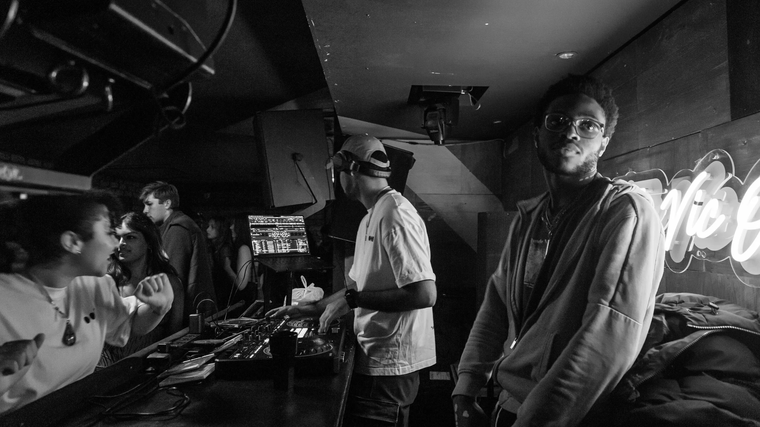 A black and white photo of a DJ mixing music at a club with a crowd of people dancing and socializing around him.