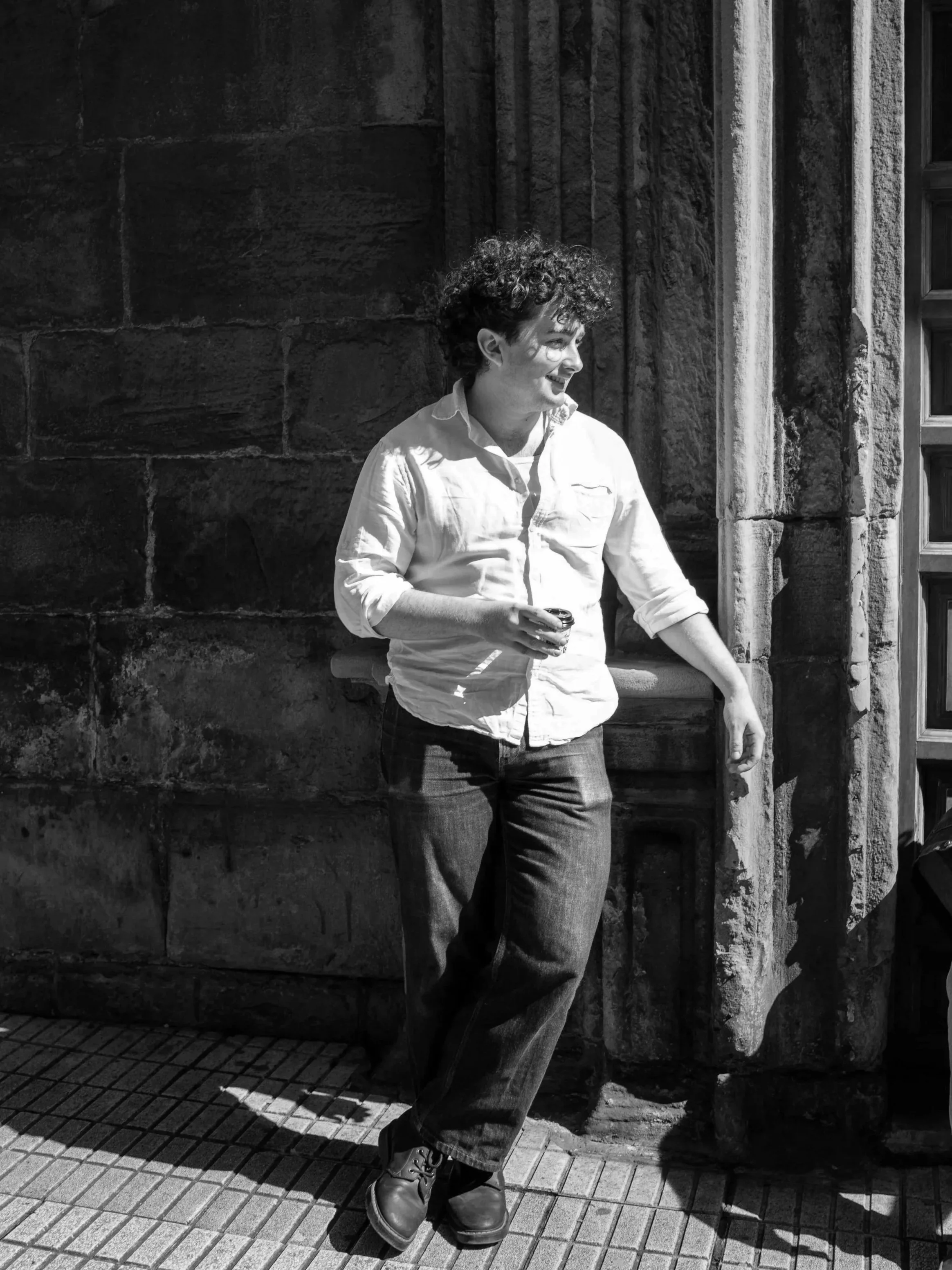 A person with curly hair wearing a white shirt and jeans standing against a stone wall, holding an object in their right hand, smiling, and looking to the right.