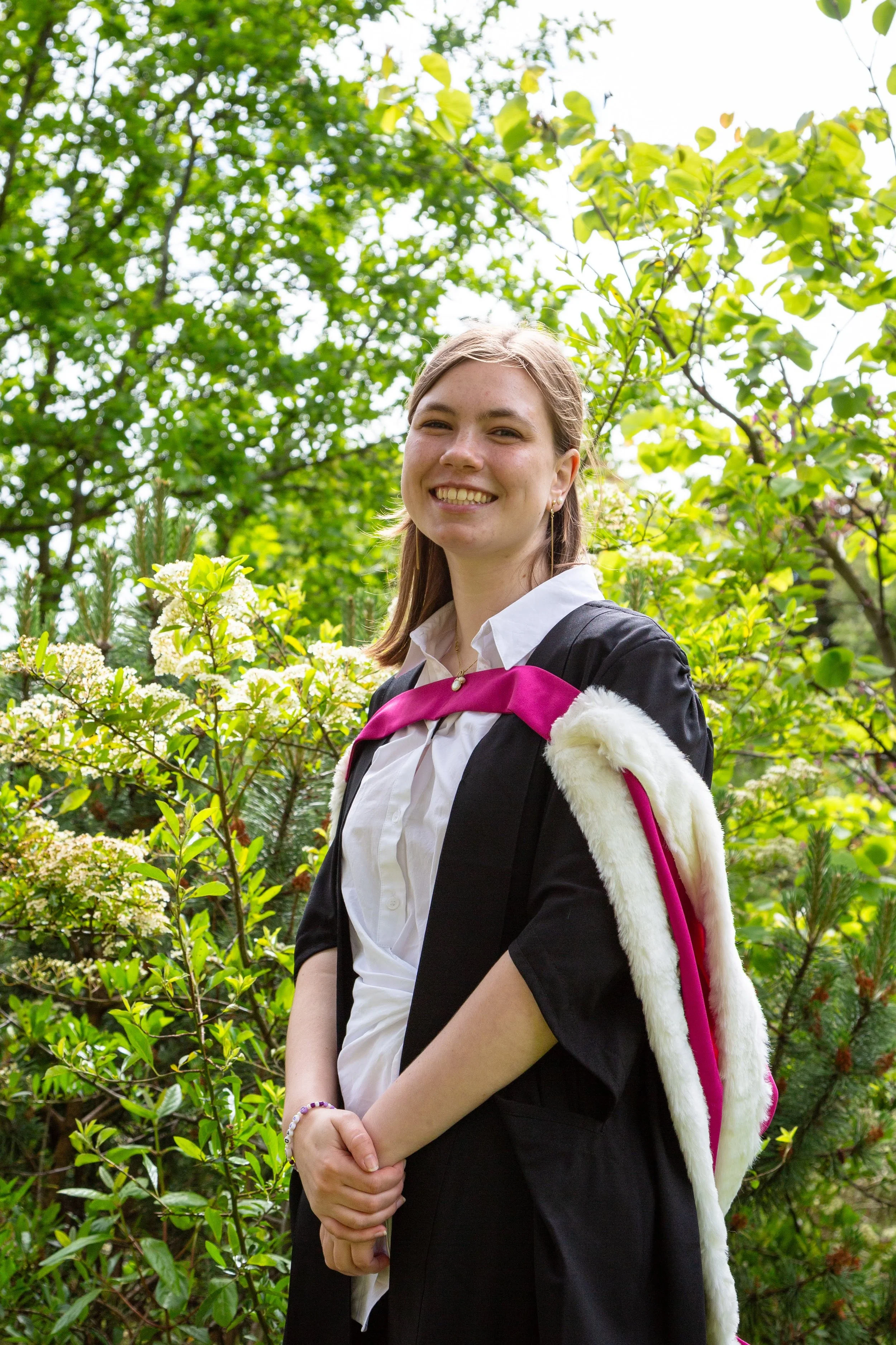 A young woman smiling outdoors amongst green trees and blooming bushes, wearing a black blazer, white shirt, and a pink backpack with white fur accents.