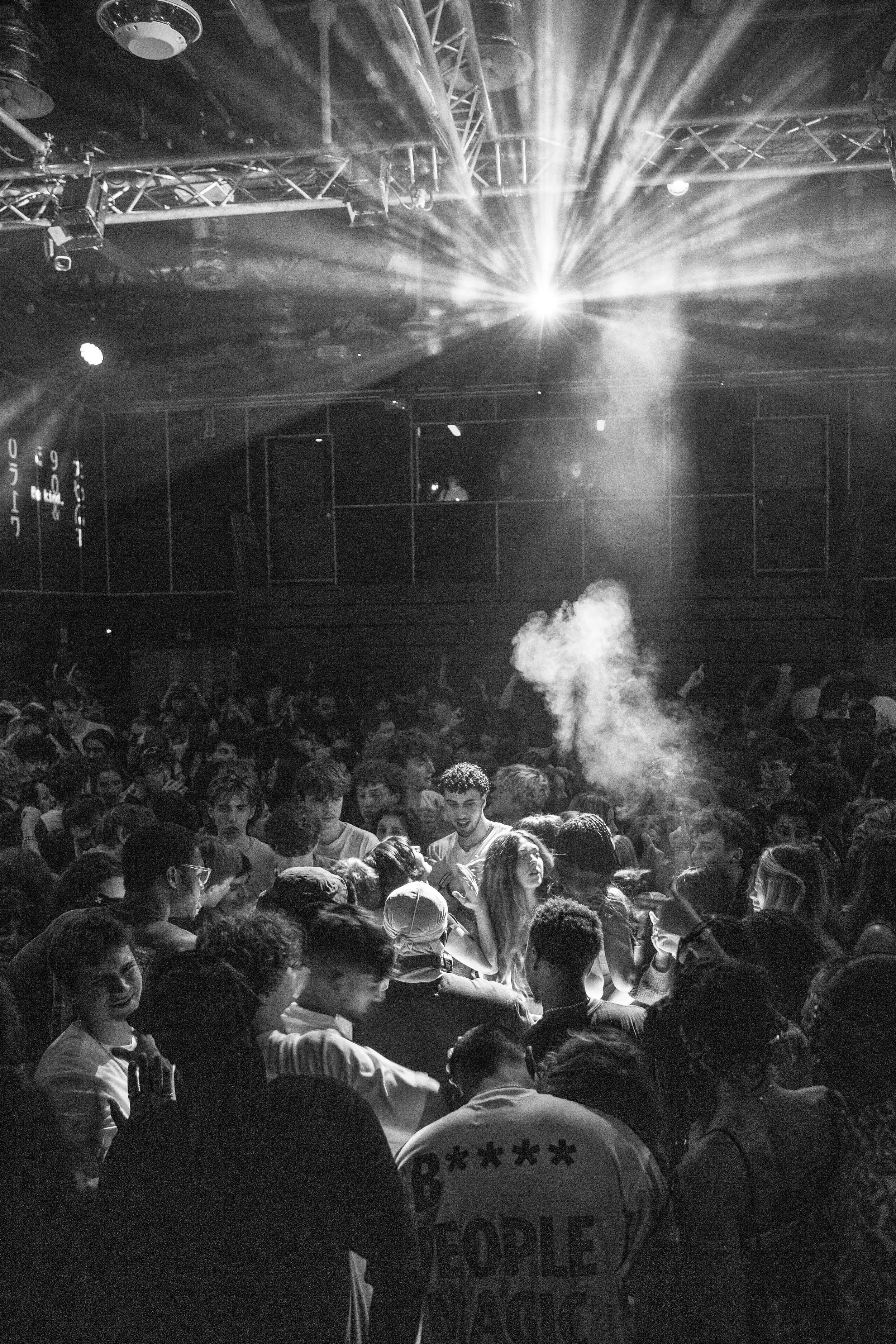 A crowded dance floor with many young people dancing and socializing, illuminated by stage lights and fog effects, suggesting a concert or nightclub setting.