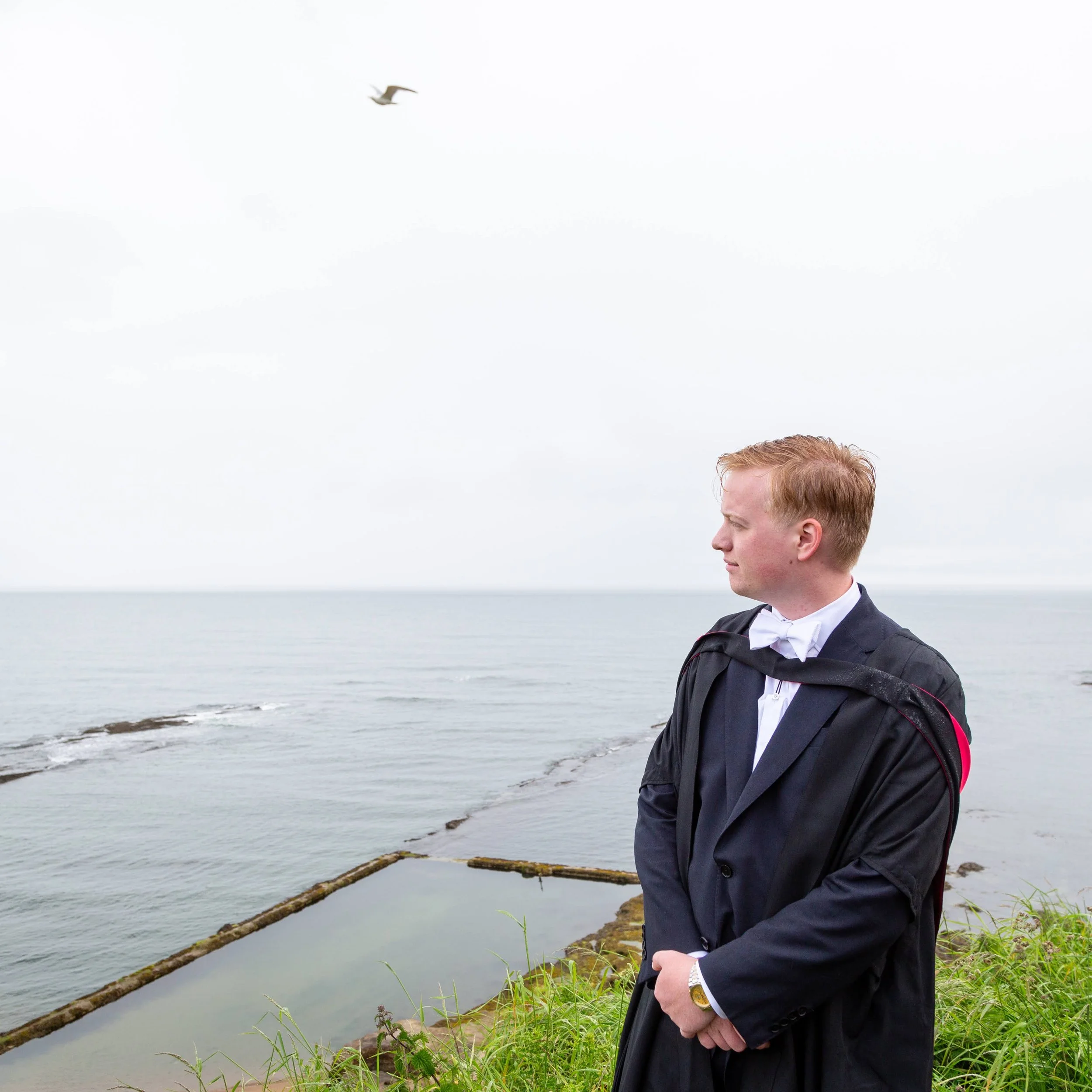 A man in a suit and tie stands by the edge of a body of water with grass and rocks, looking to the side with a calm expression, with the ocean and sky in the background.