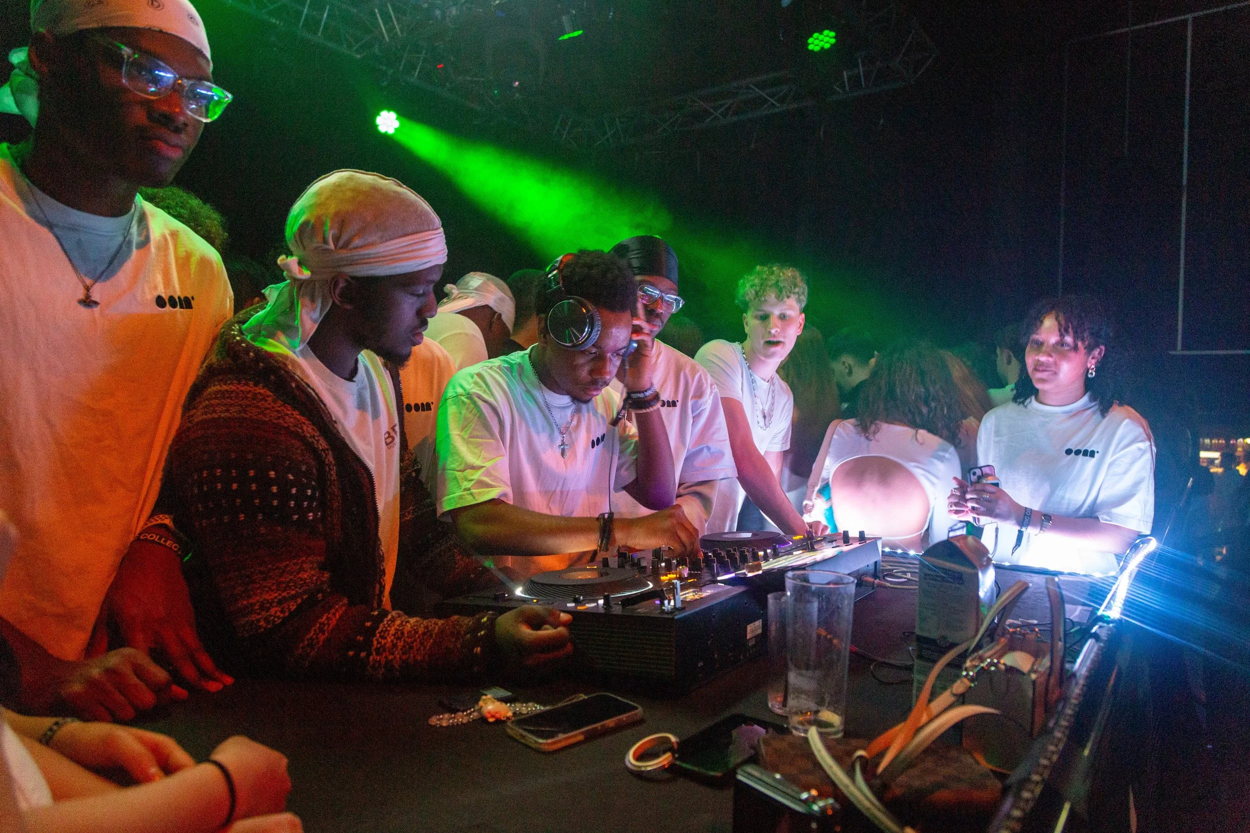 Group of young people in a dark indoor setting with green lighting, some wearing headphones, gathered around DJ equipment, enjoying a music event or party.
