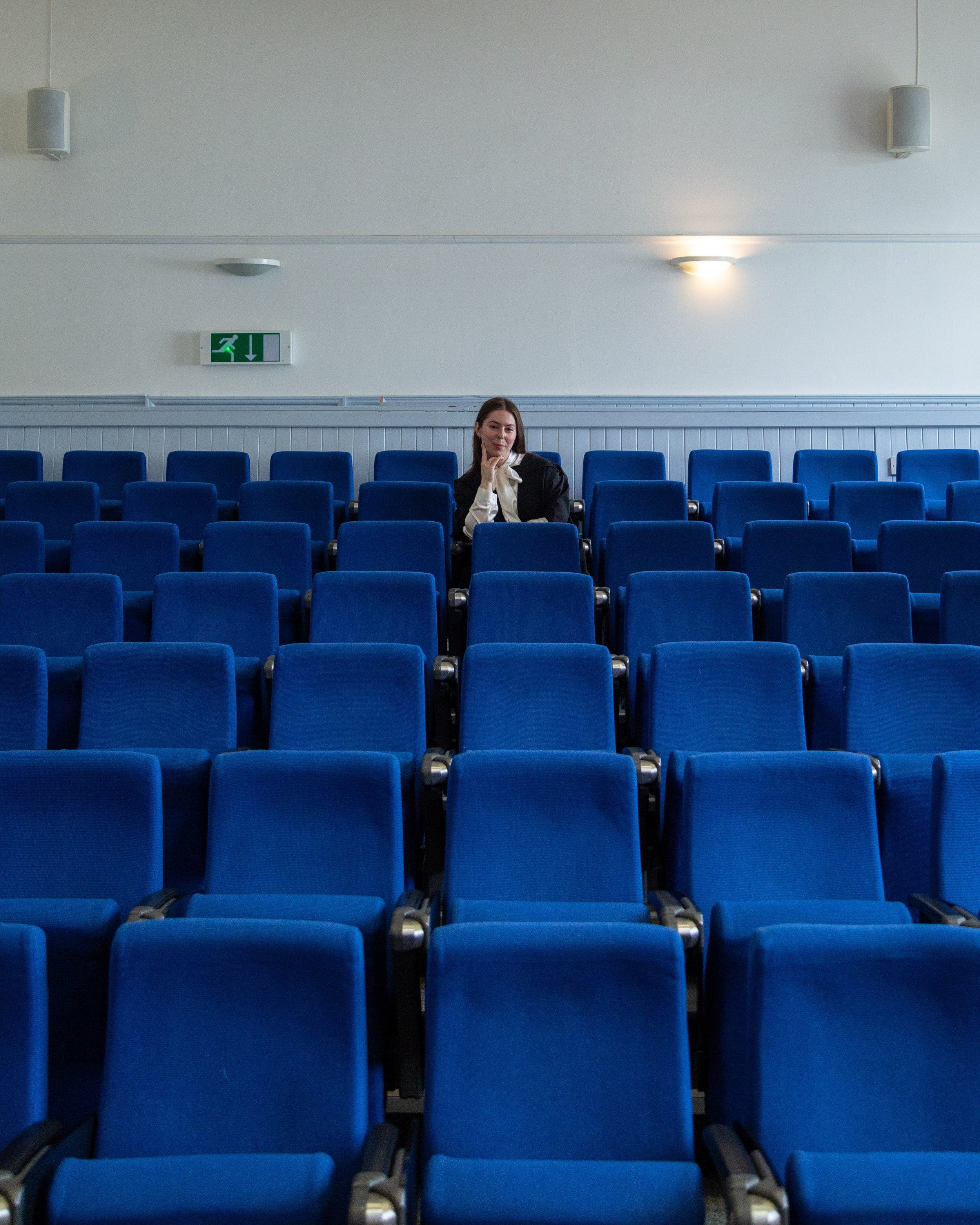 An empty auditorium with rows of blue chairs and a woman sitting in the middle row, resting her chin on her hand, with an emergency exit sign above her on the wall.