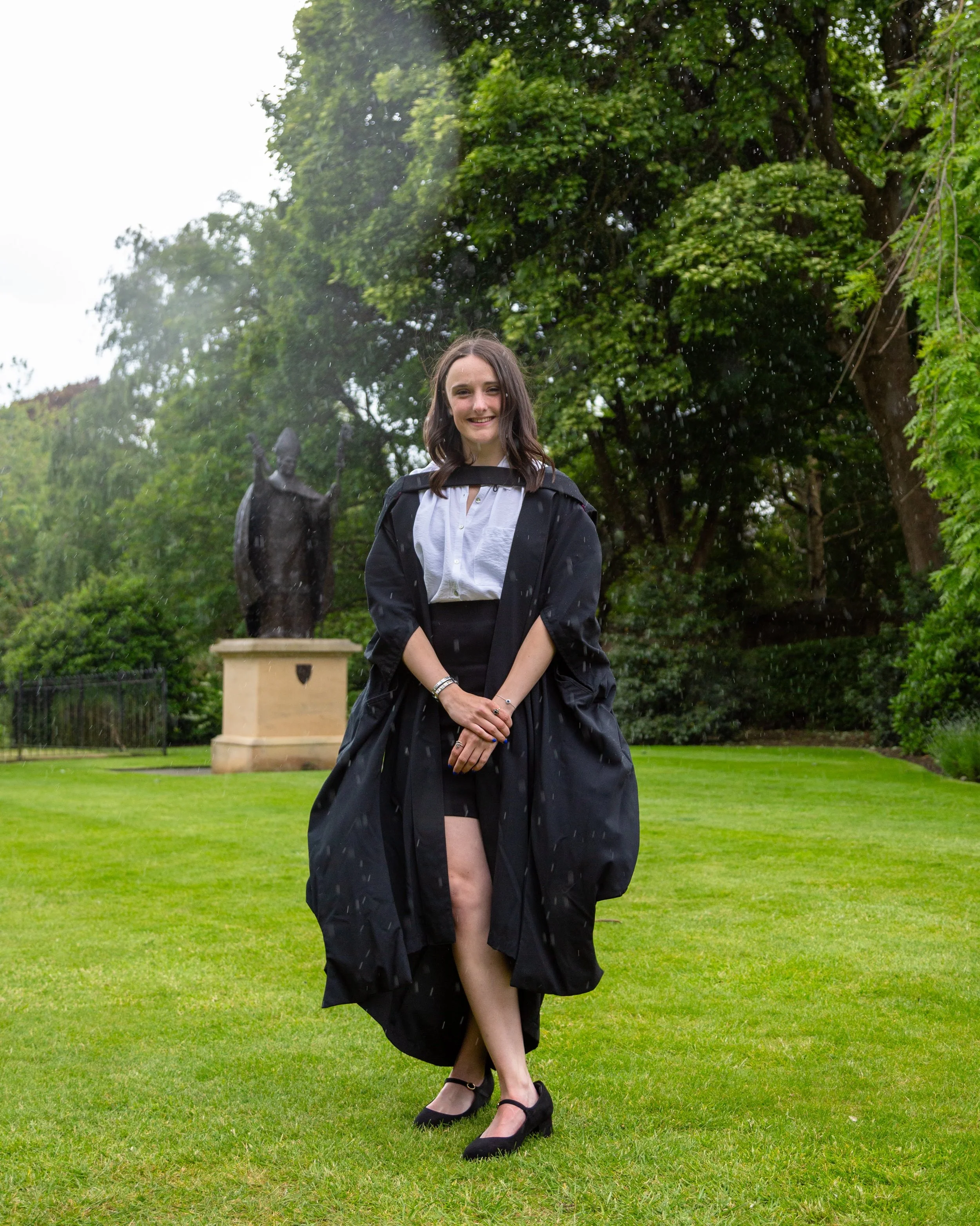 Young woman with dark brown hair wearing a black gown and heels, standing on a lush green lawn with a statue and large trees in the background on a rainy day.