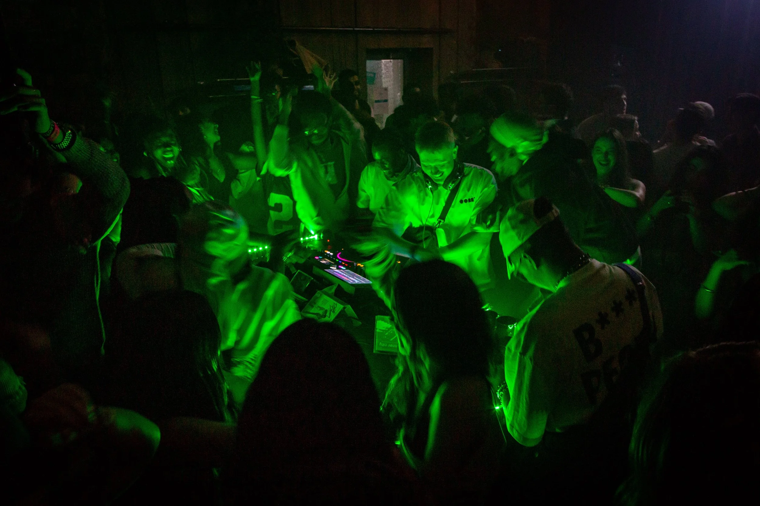 People dancing and a DJ in a nightclub with a dark interior and neon sign.