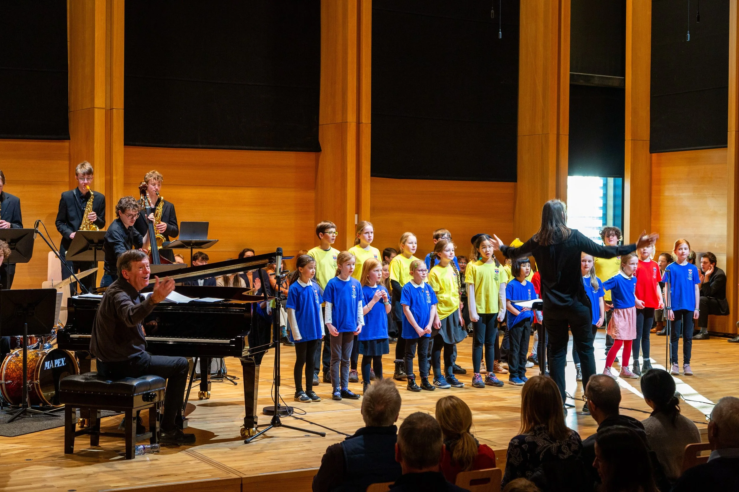 Youth choir performing on stage with conductor and musicians, in a wooden auditorium with audience members watching.