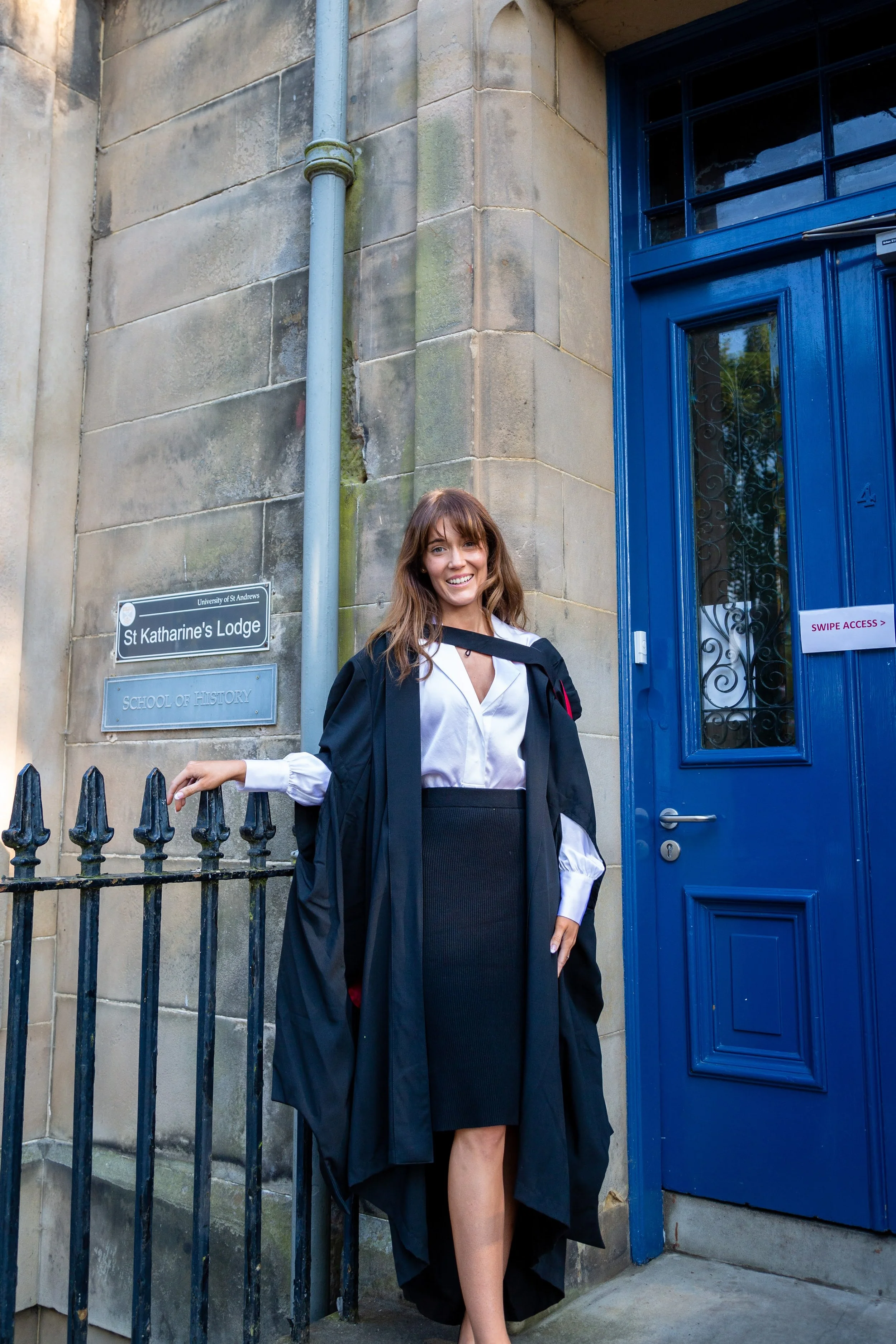 A young woman in a graduation cap and gown smiling and standing outside a stone building with a blue door and signs that read 'St Katharine's Lodge' and 'School of History'.