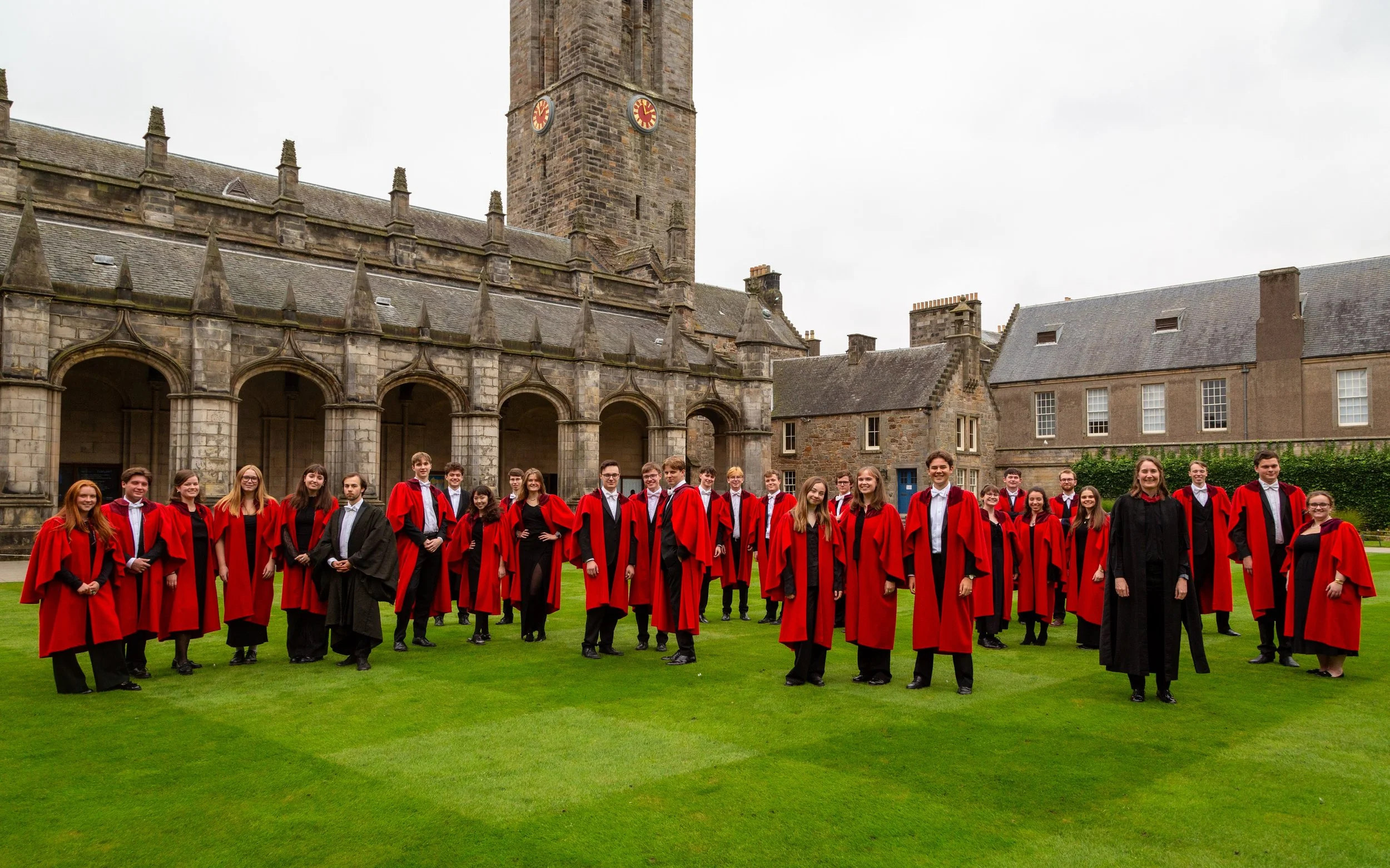 A group of students in graduation robes standing on a green lawn in front of a historic stone building with a clock tower.