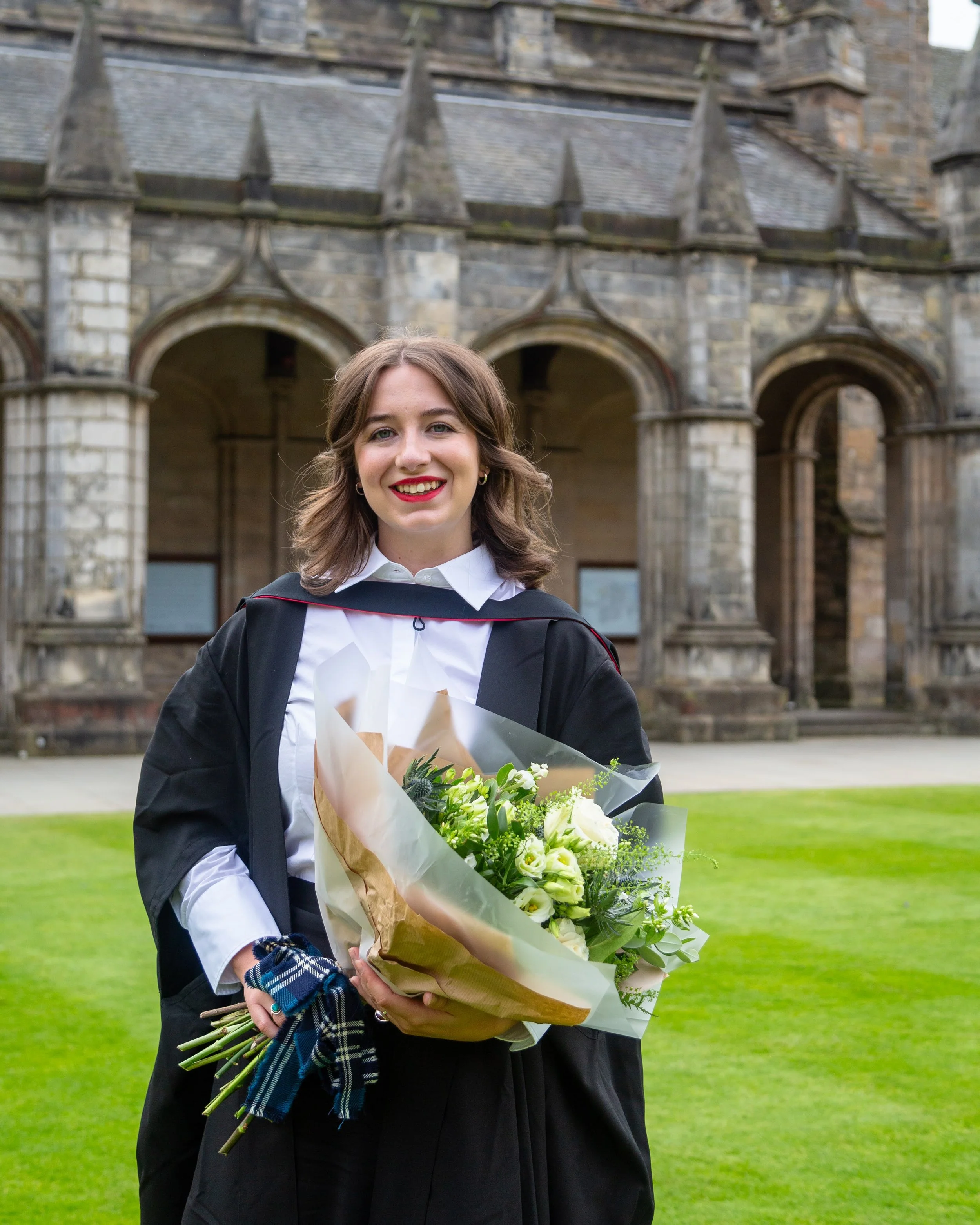 A young woman wearing a graduation gown and holding a bouquet of white flowers, standing outdoors in front of a historic stone building with arched windows and pointed turrets.