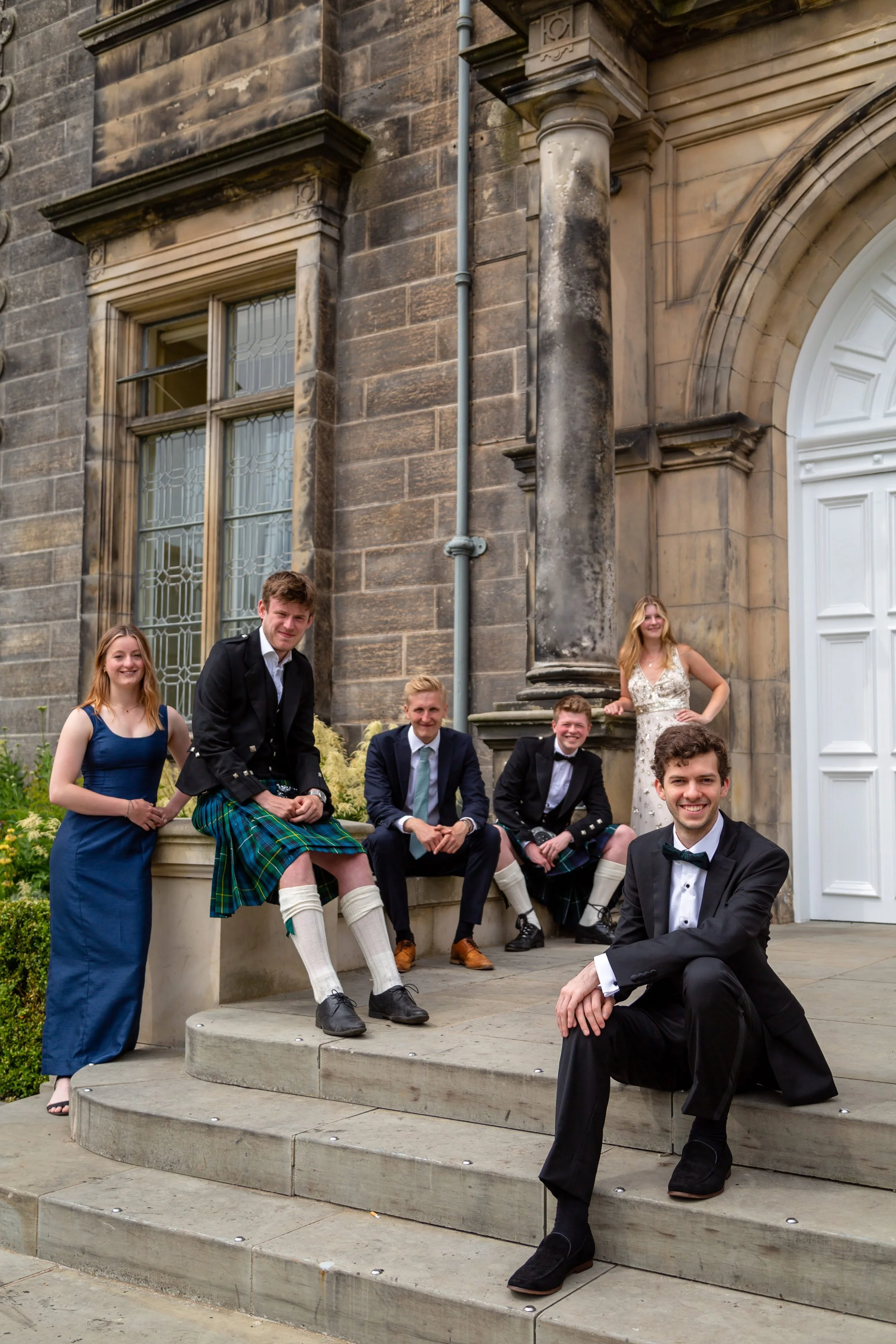 Group of young adults dressed in formal and traditional Scottish attire, posing on the steps outside a historic building with large windows and stone columns.