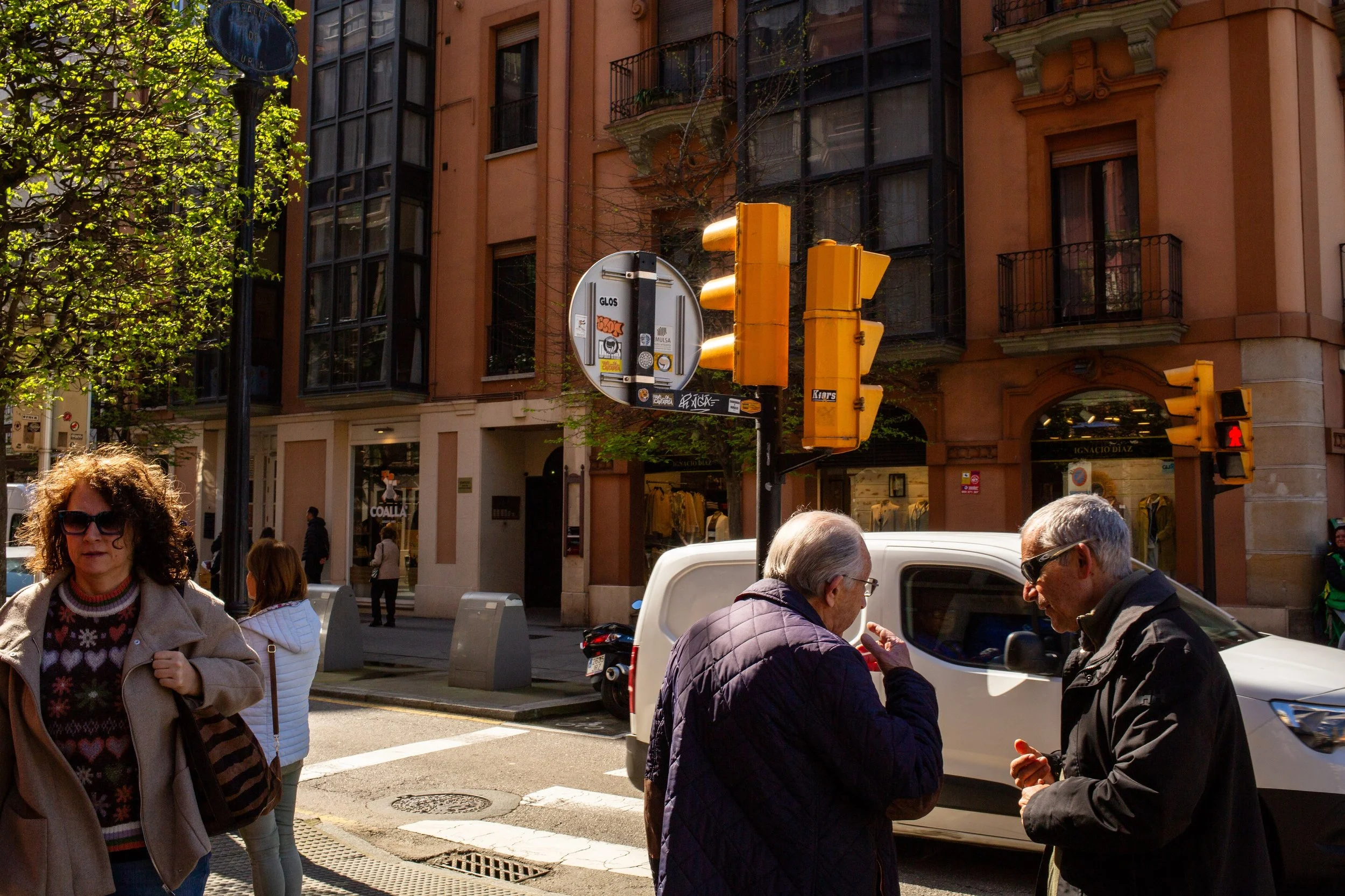 People crossing the street in front of a building with traffic lights and street signs, some looking at their phones, some walking, in an urban area.