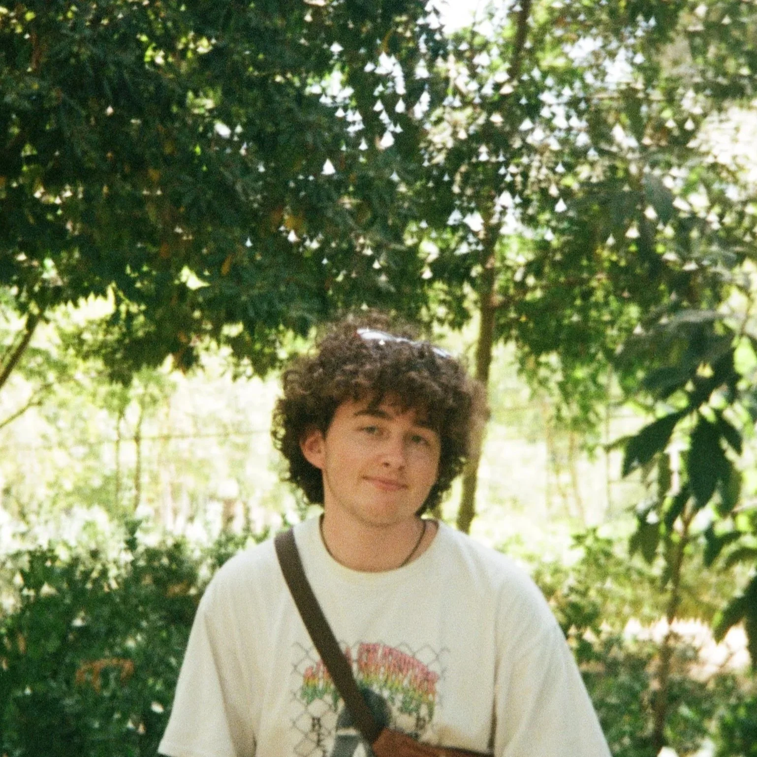 A young man with curly hair standing outdoors in front of green trees, wearing a white T-shirt and carrying a shoulder bag.