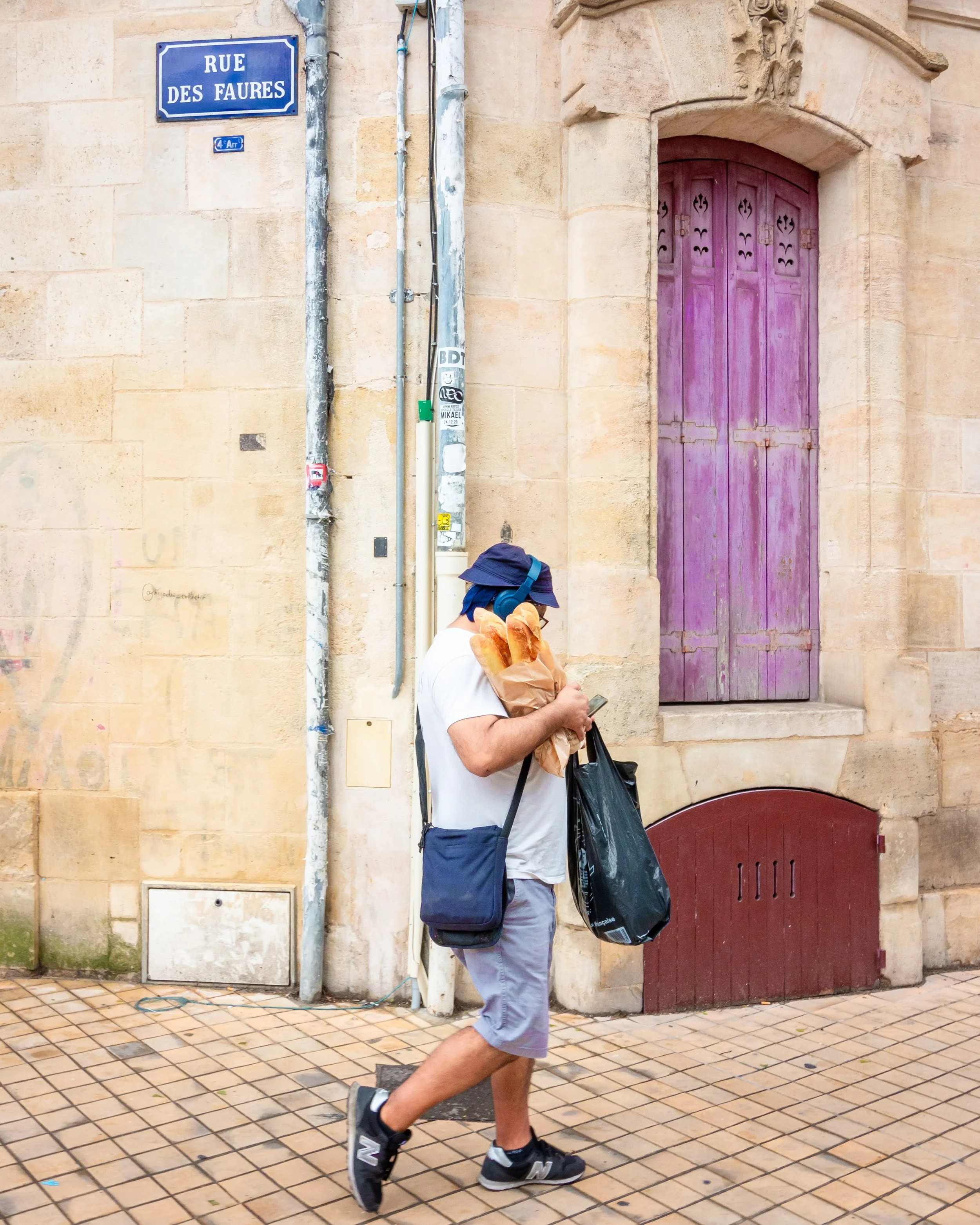 A person walking on a tiled sidewalk holding a paper bag with baguettes and a phone, wearing a hat and headphones, with a beige stone building in the background featuring a purple wooden door and street signs reading 'Rue des Faures' and '4th arrondi