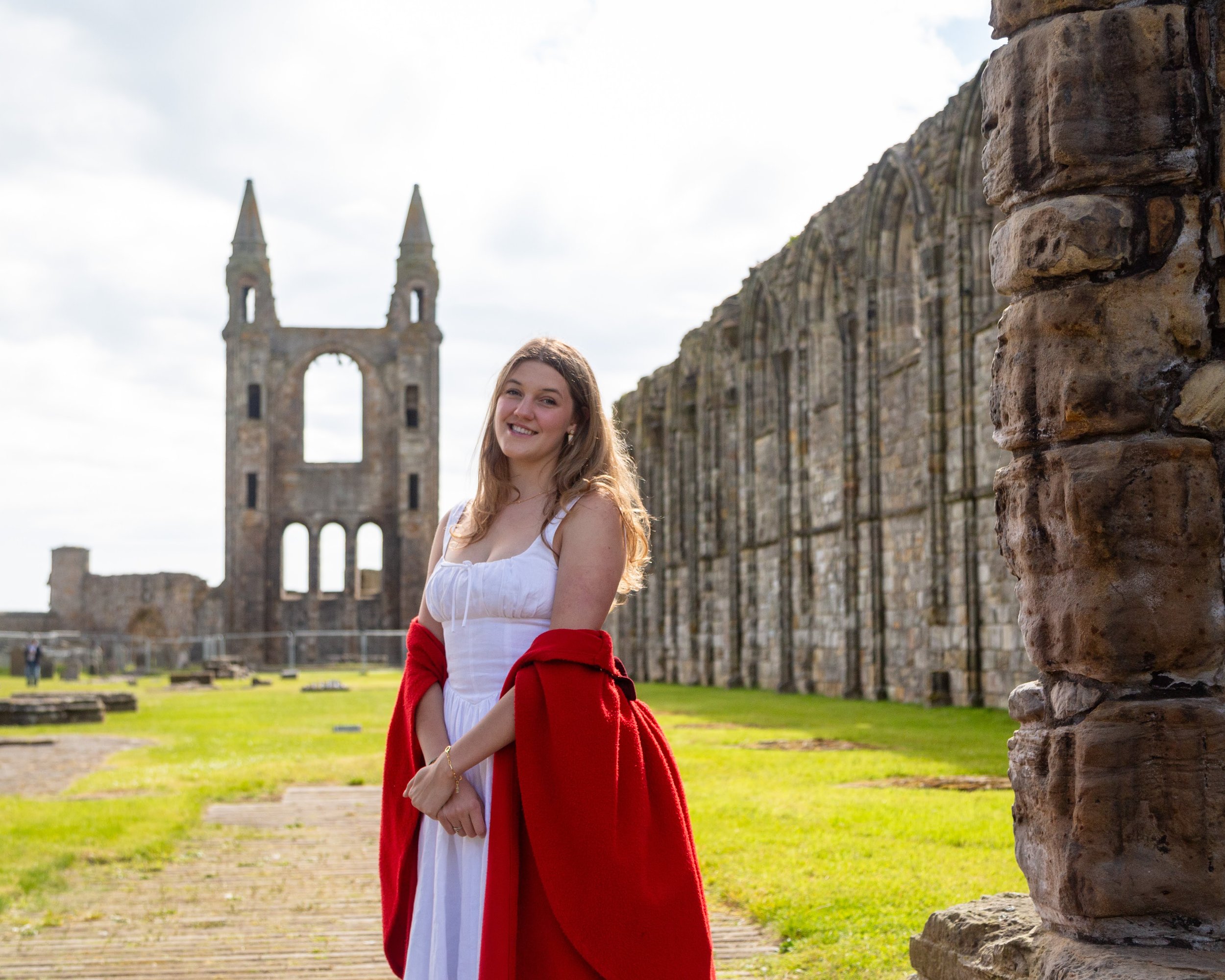 A young woman with long wavy hair smiling at the camera, standing outdoors near a stone wall with ruins in the background, wearing a white dress with a red jacket draped over her shoulders.