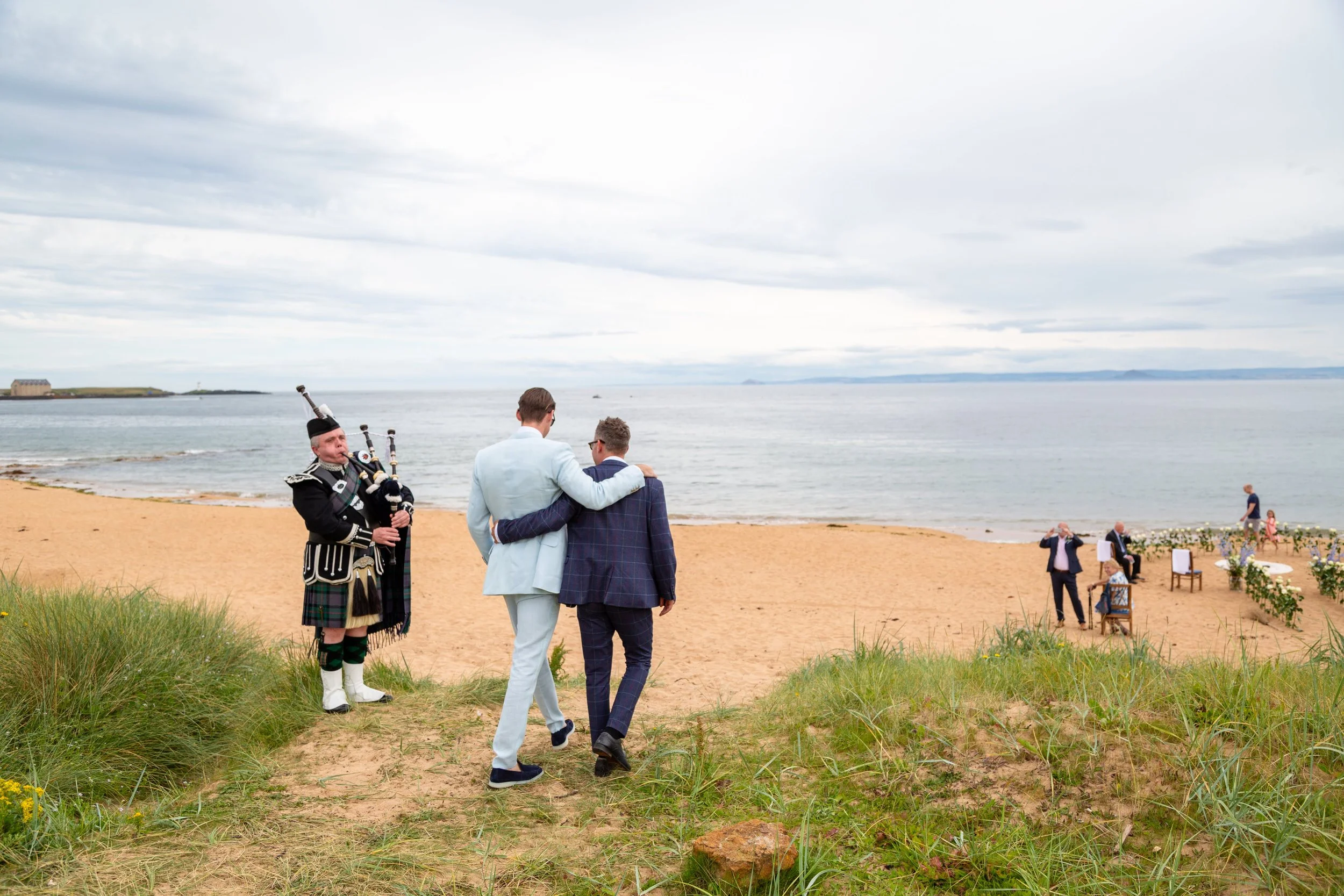 A wedding ceremony on the beach with two men in suits walking hand in hand, a bagpiper in traditional Scottish attire playing, and guests seated nearby, overlooking calm ocean waters under a cloudy sky.