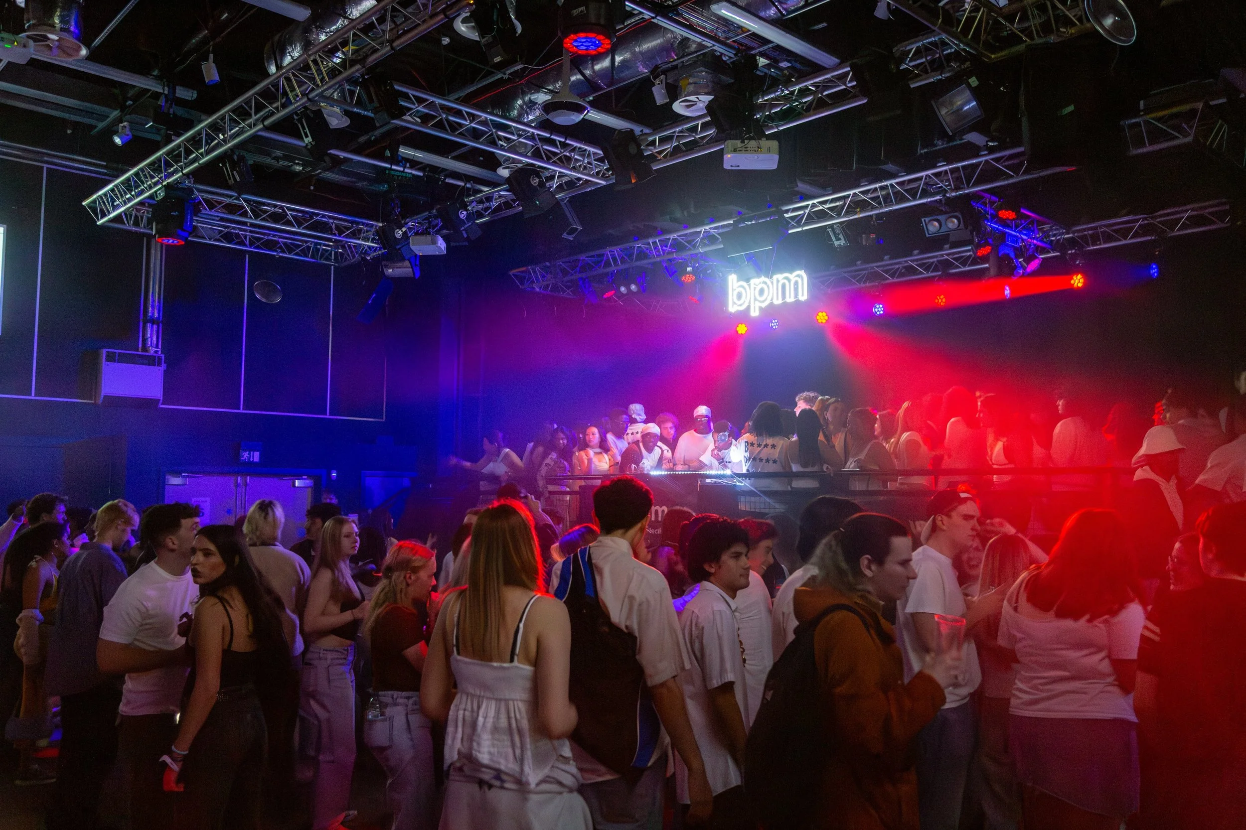 A crowded nightclub with people dancing and socializing under colorful lighting. The stage has a neon sign that reads 'bpm' and various lighting fixtures and equipment above.