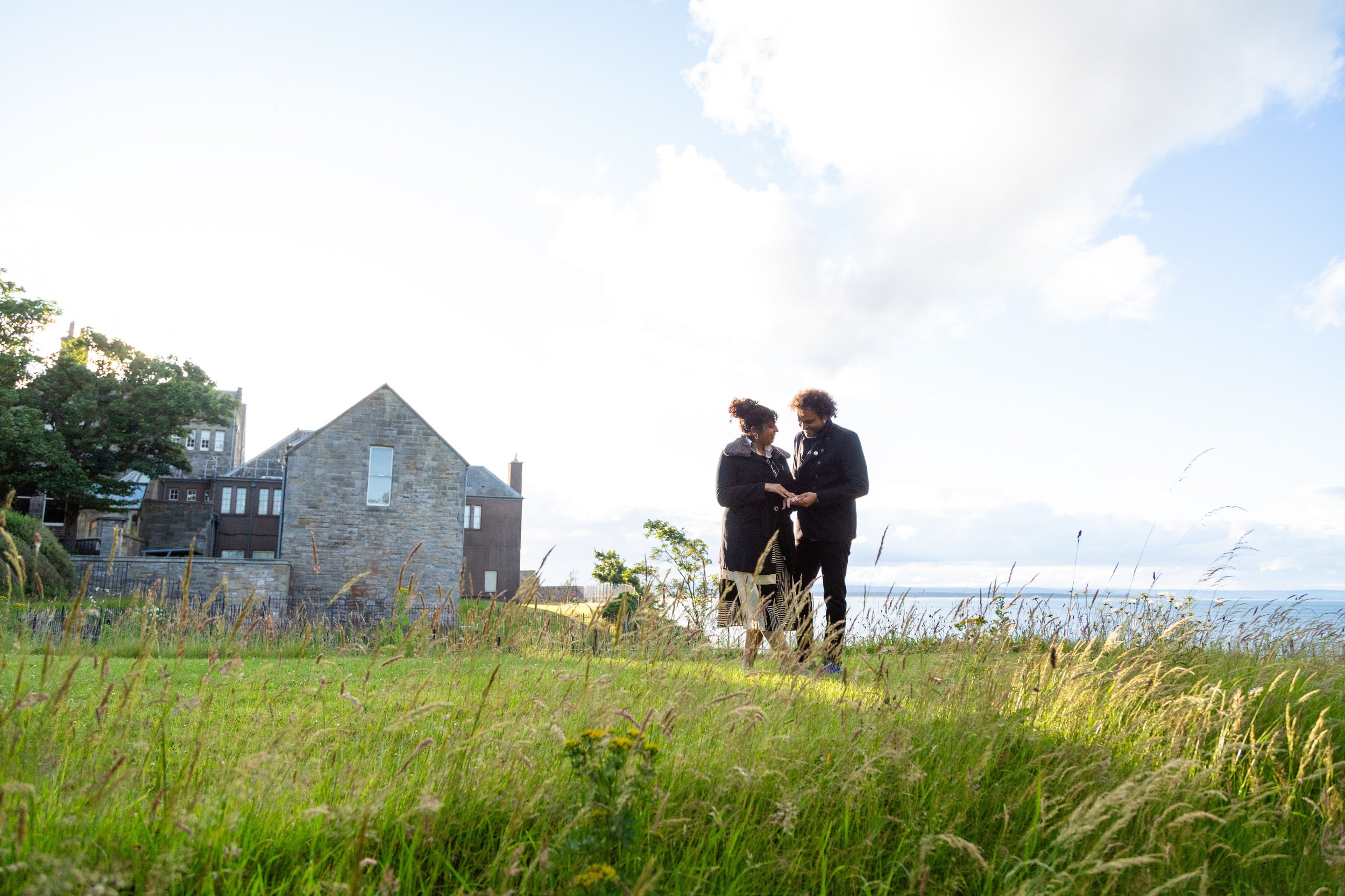 A couple standing outdoors on a grassy field, holding hands and looking at each other, with a background of an old stone house, trees, and a cloudy sky.