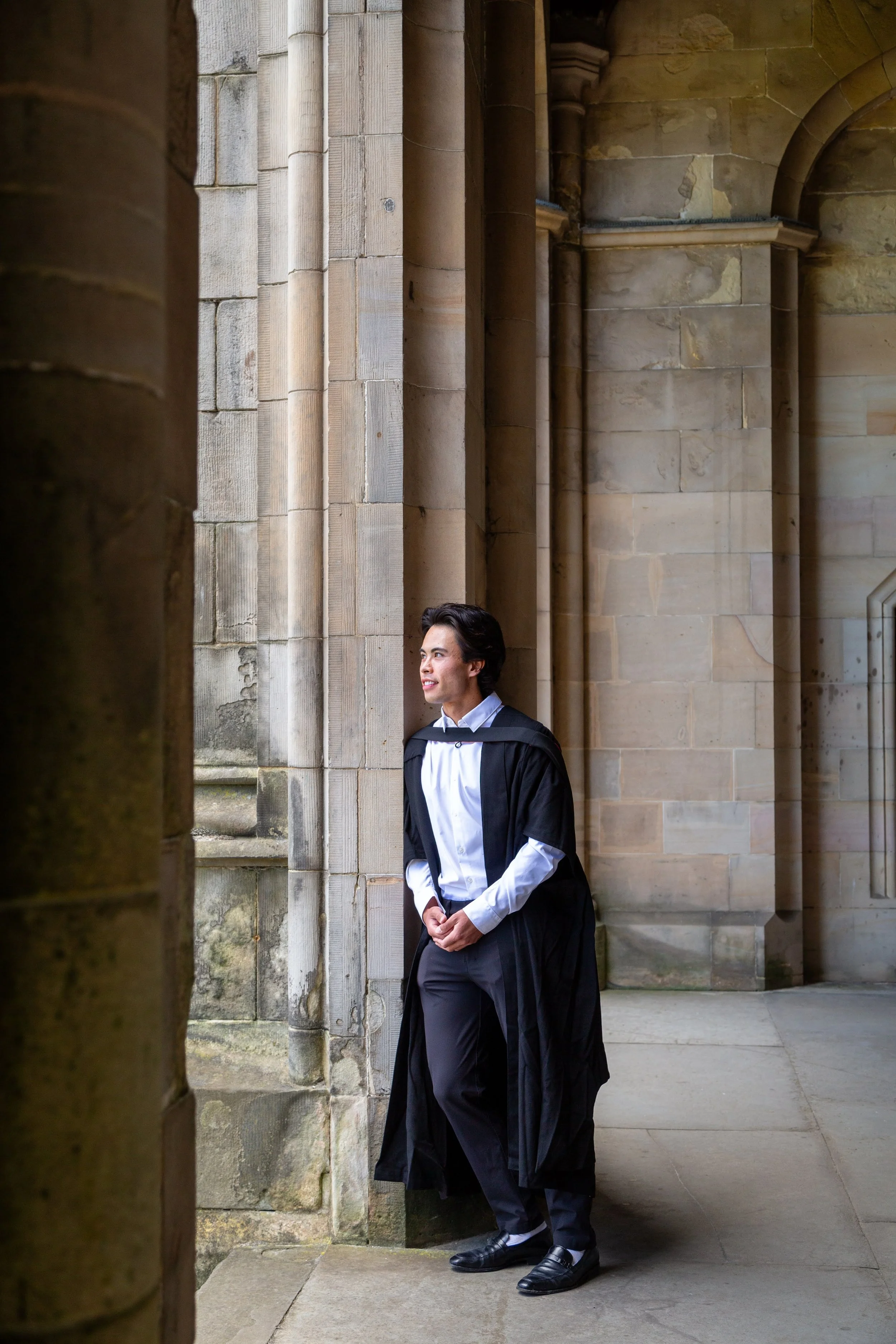 Young man dressed in formal attire, standing quietly in a stone corridor with tall columns, possibly in a historic or academic building.