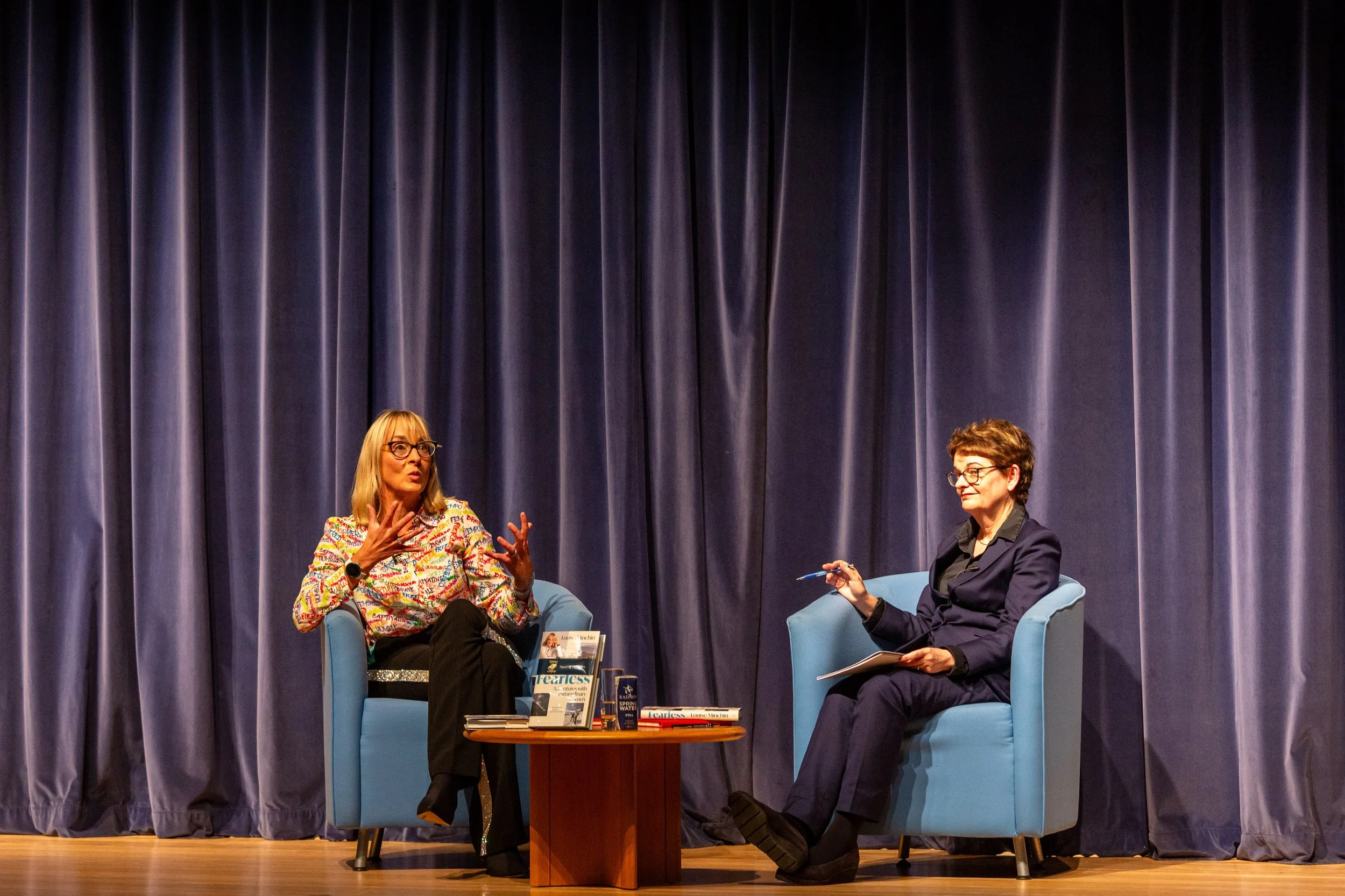 Two women engaged in conversation on stage with blue curtains in the background. One woman wears glasses and a colorful patterned shirt, gesturing with her hands. The other woman, also wearing glasses, is dressed in a dark blazer and holds a pen and notepad. A small table with books and brochures is between them, and they sit in light blue armchairs.