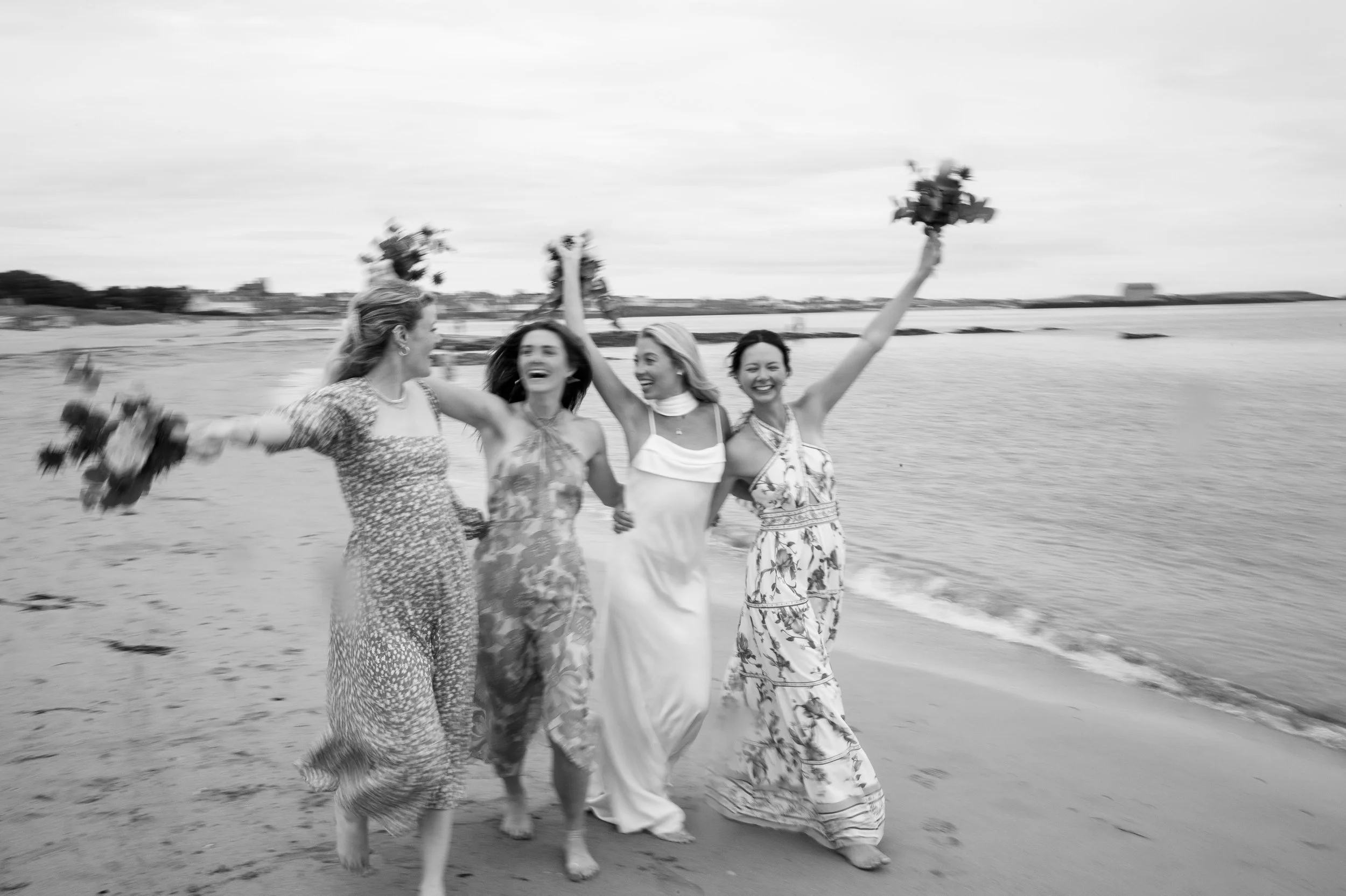 Four women in dresses walking along a beach, smiling and holding bouquets, with two women raising bouquets in the air, beach and water in background.