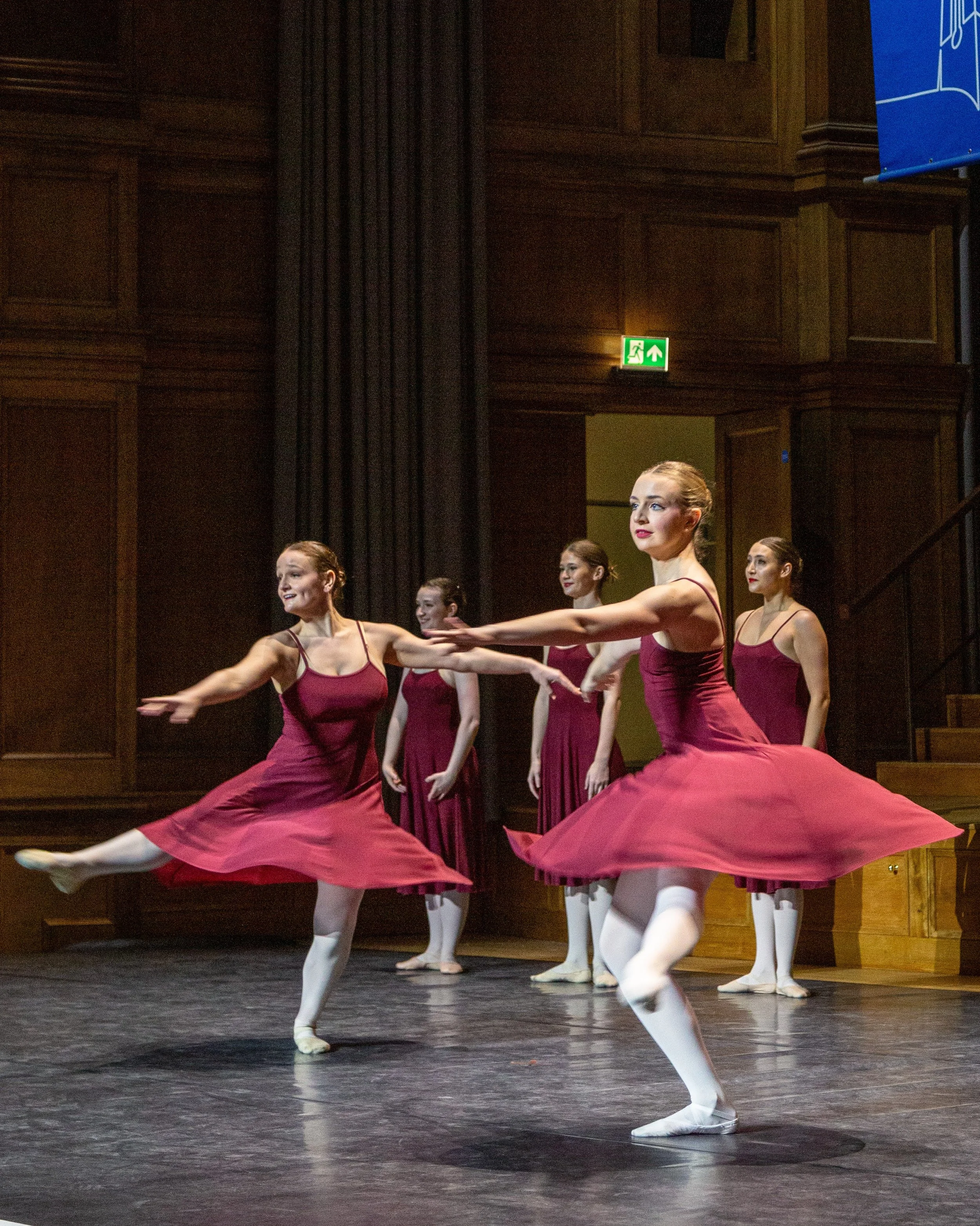 A group of ballet dancers wearing maroon tutus and white tights practicing in a wooden stage with dark curtains and wood-paneled walls, with one dancer in front extending her arms and legs gracefully.