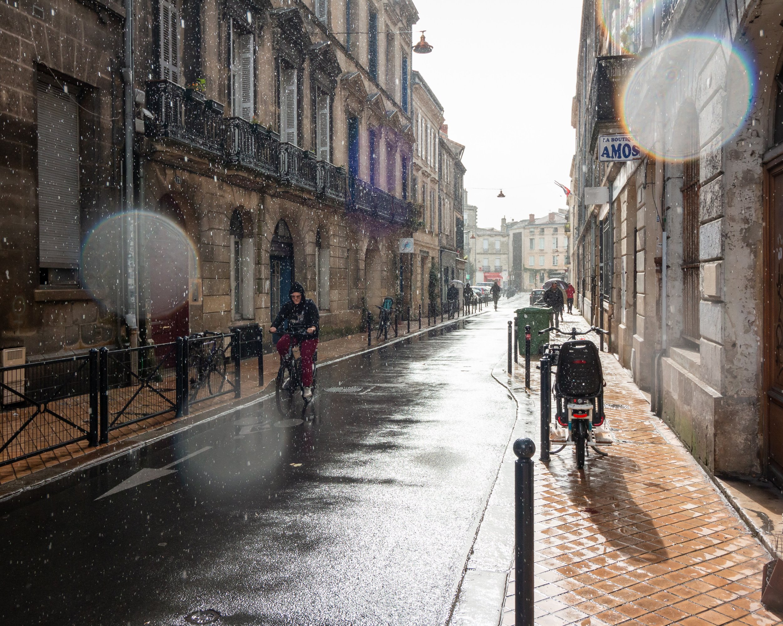 A city street scene after rain with sunlight breaking through. Wet pavement with reflections, buildings with balconies, and several people walking or riding bikes under umbrellas.