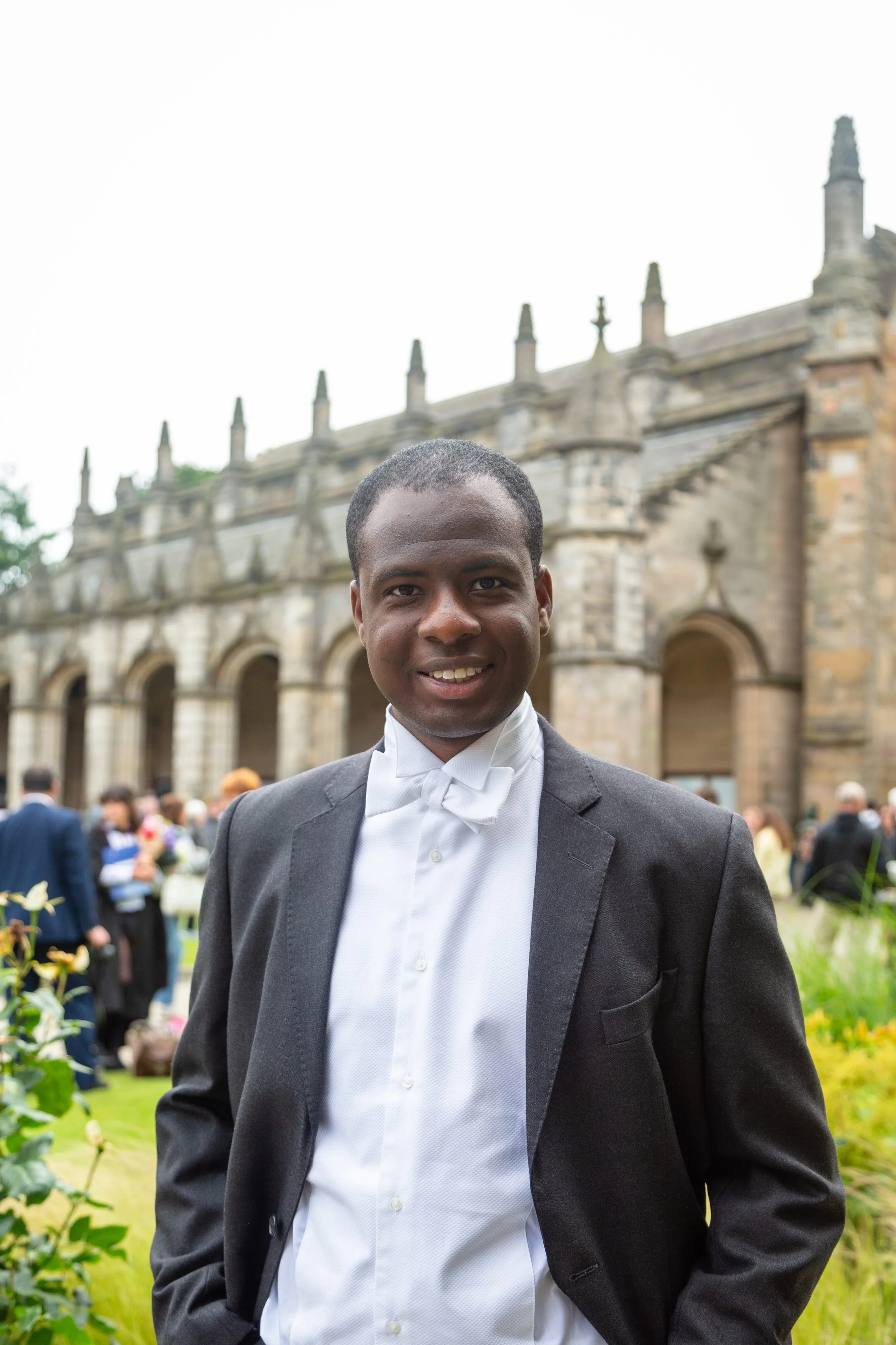 A young man in a tuxedo standing outdoors in front of a historic stone building with Gothic architectural features, surrounded by a group of people at a formal event.