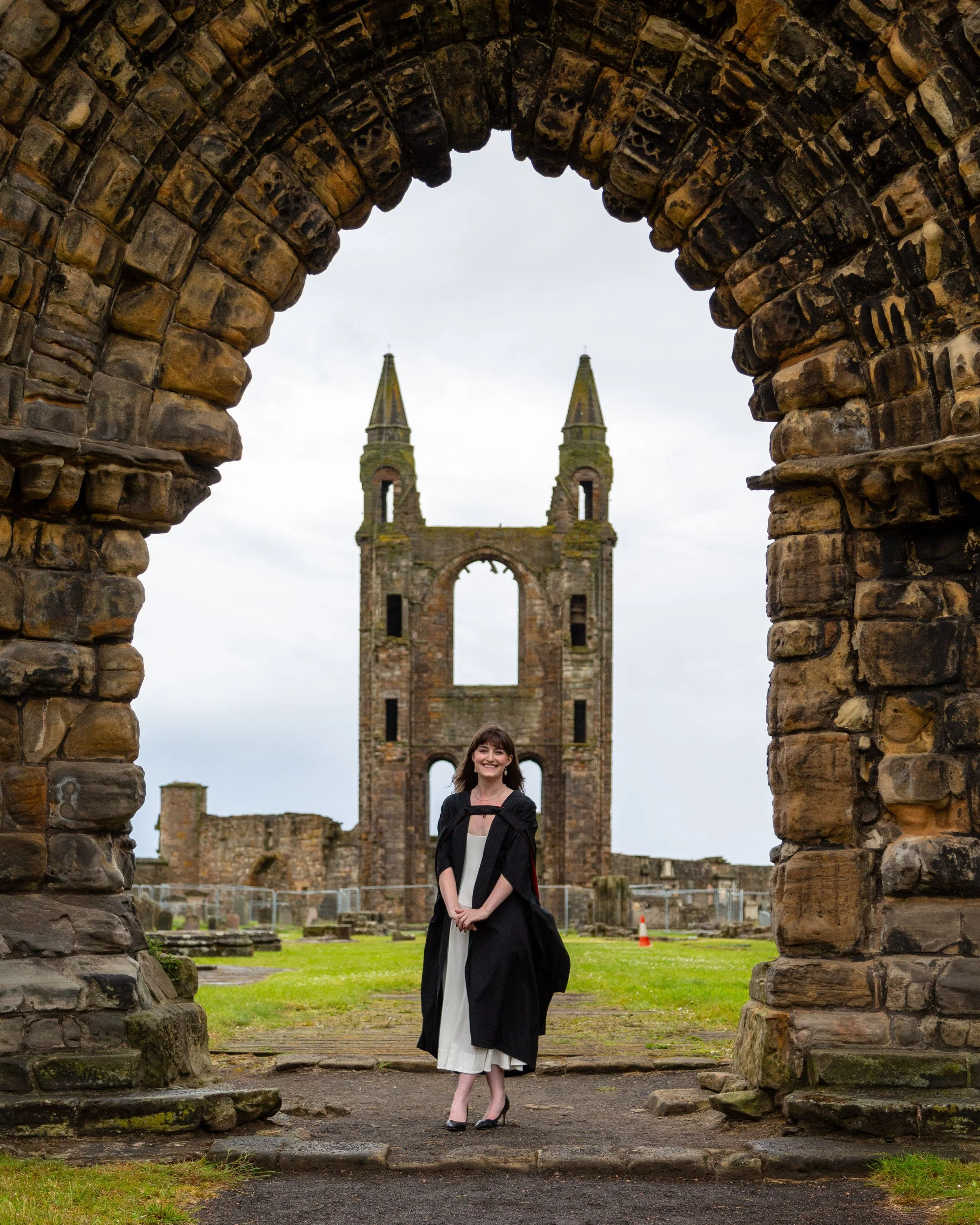 Young woman standing in historical stone archway ruins with daylight sky and church or castle in the background.