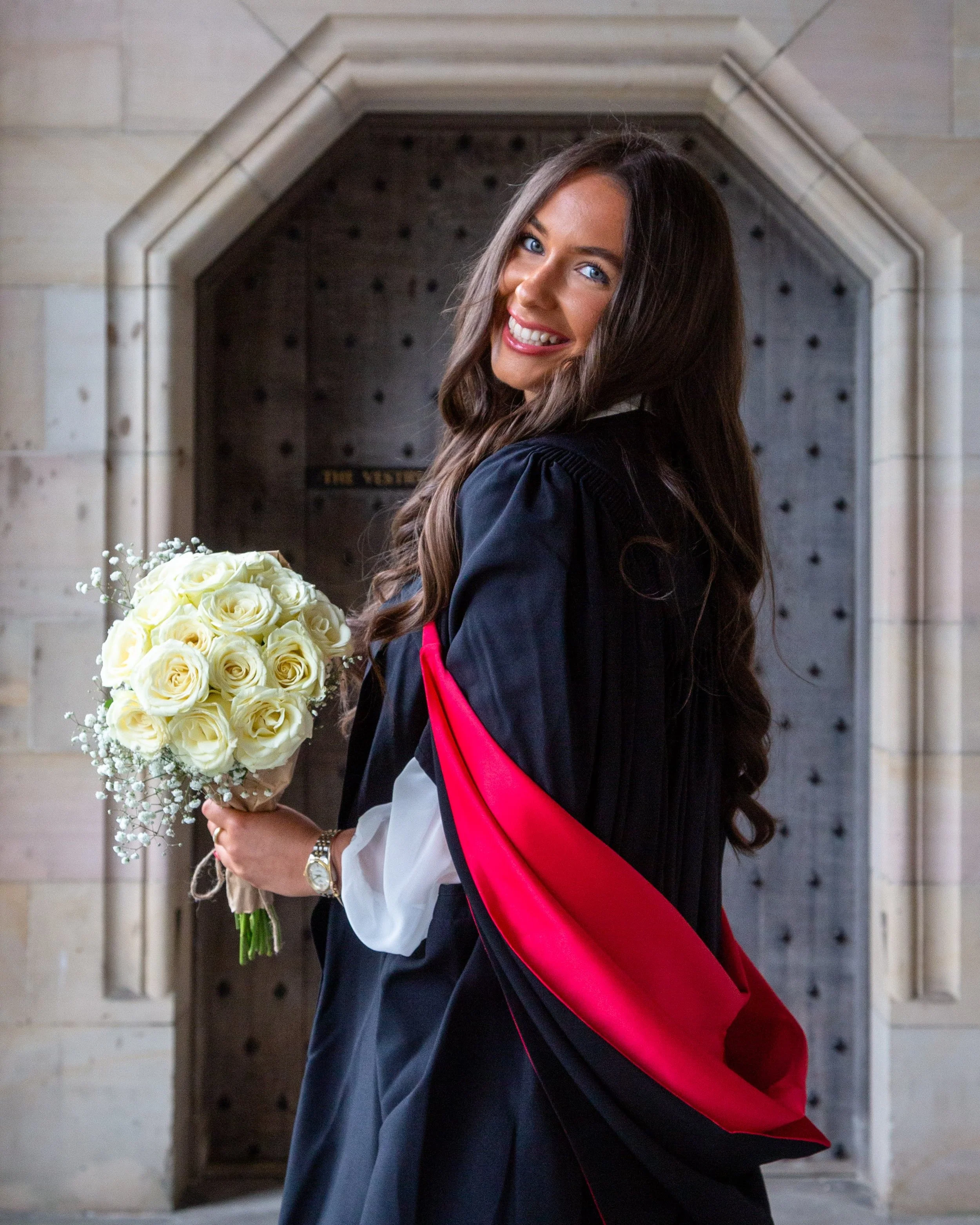 A woman with long brown hair, blue eyes, and a bright smile holding a bouquet of white roses, wearing a dark academic gown and a red stole, standing in front of an arched stone doorway.