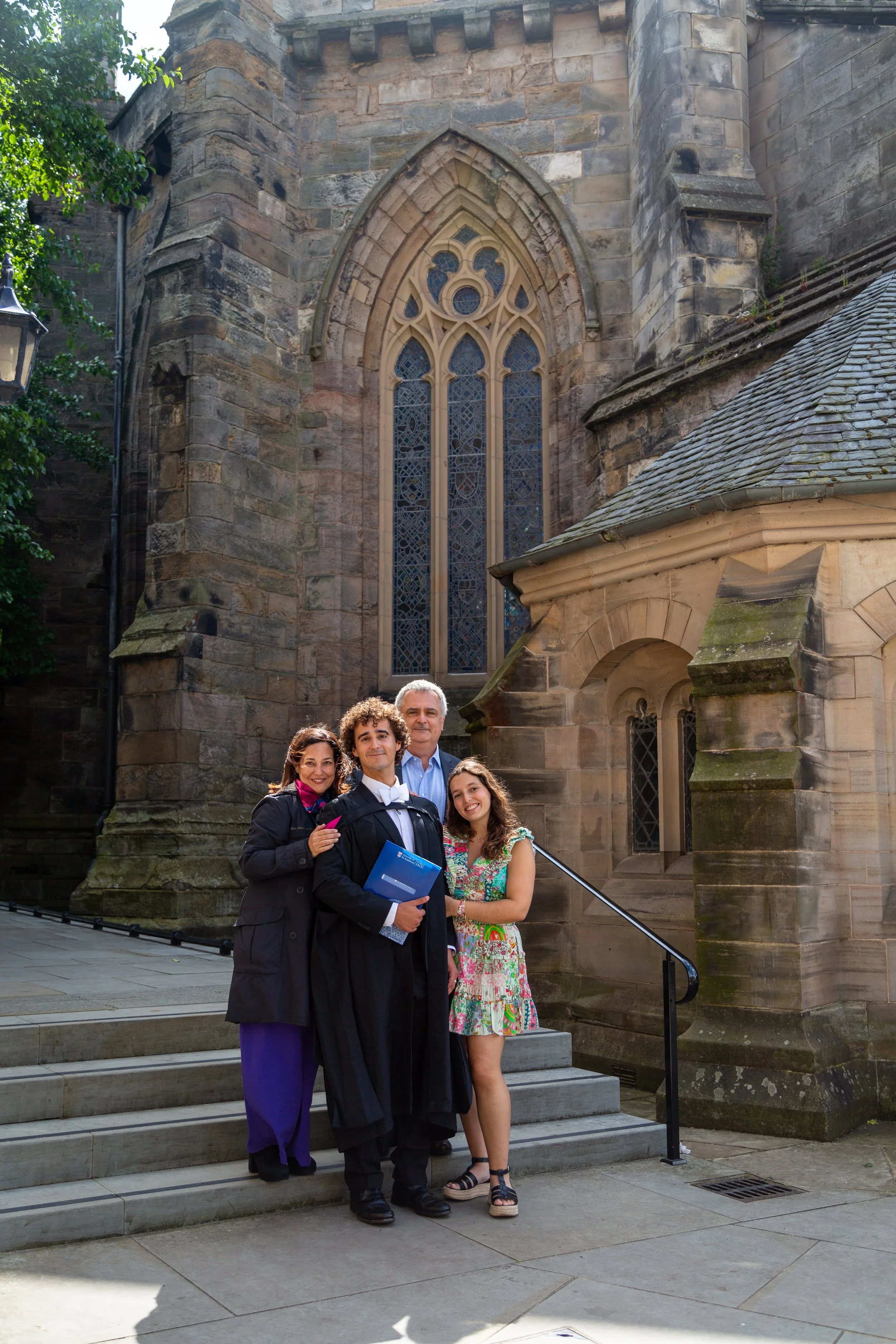 Group of five people, including a graduate in a cap and gown, standing on steps in front of a historic church or cathedral with large stained glass windows and gothic architecture.