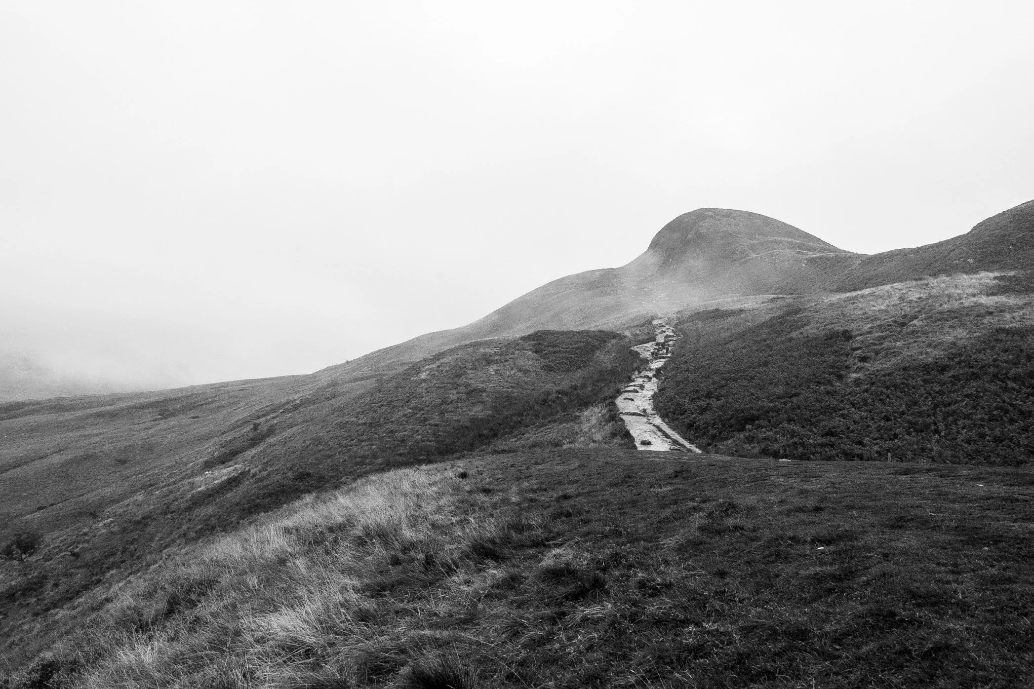 A black and white photo of a mountain landscape with a path leading up the hillside, grassy terrain in the foreground, and a rounded mountain peak in the background.