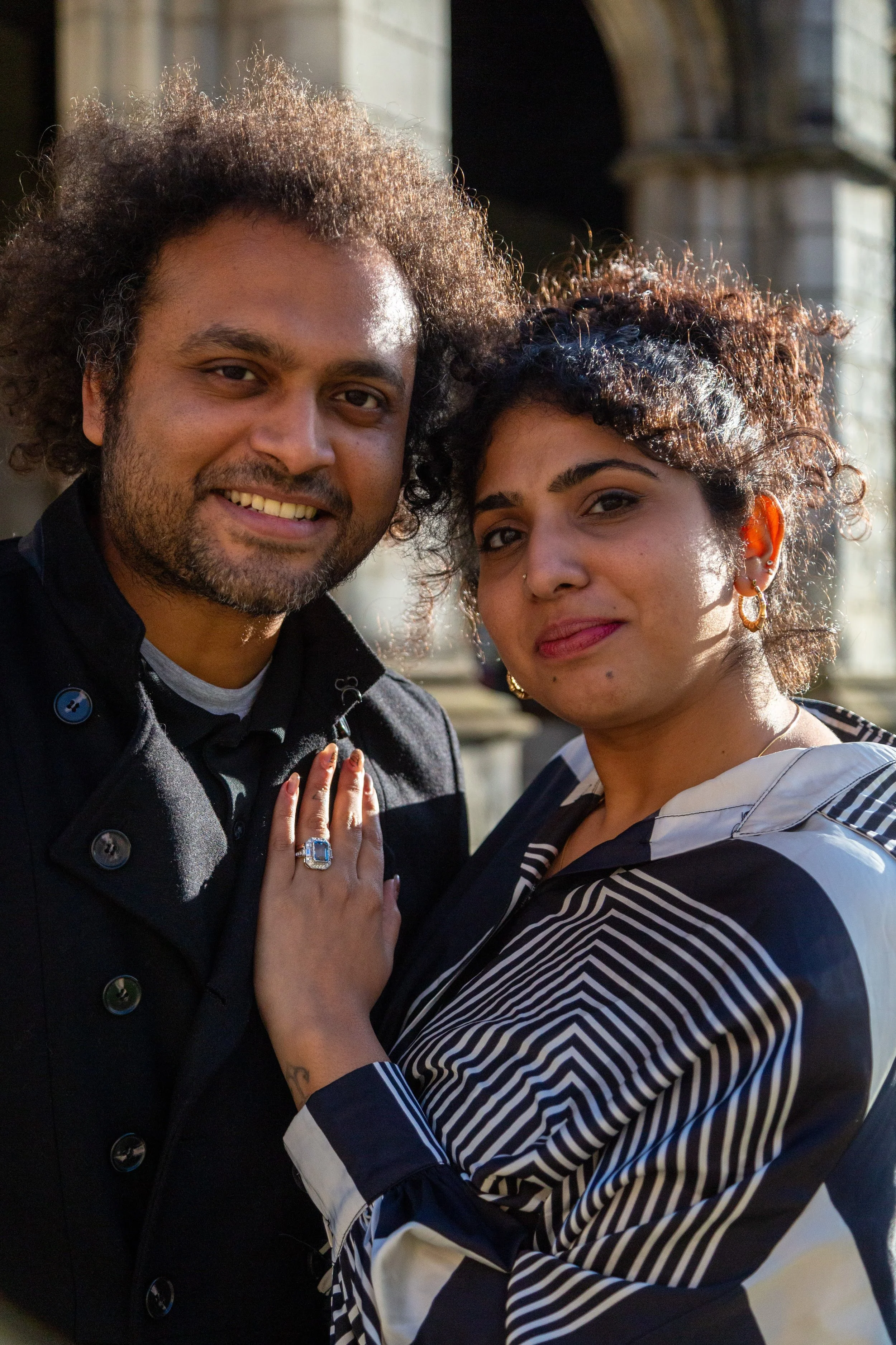 A man and a woman standing close together outdoors during daylight, smiling at the camera. The man has curly hair and facial hair, and the woman has curly hair with earrings and a nose piercing, wearing a striped blazer. Their hands are touching, wit