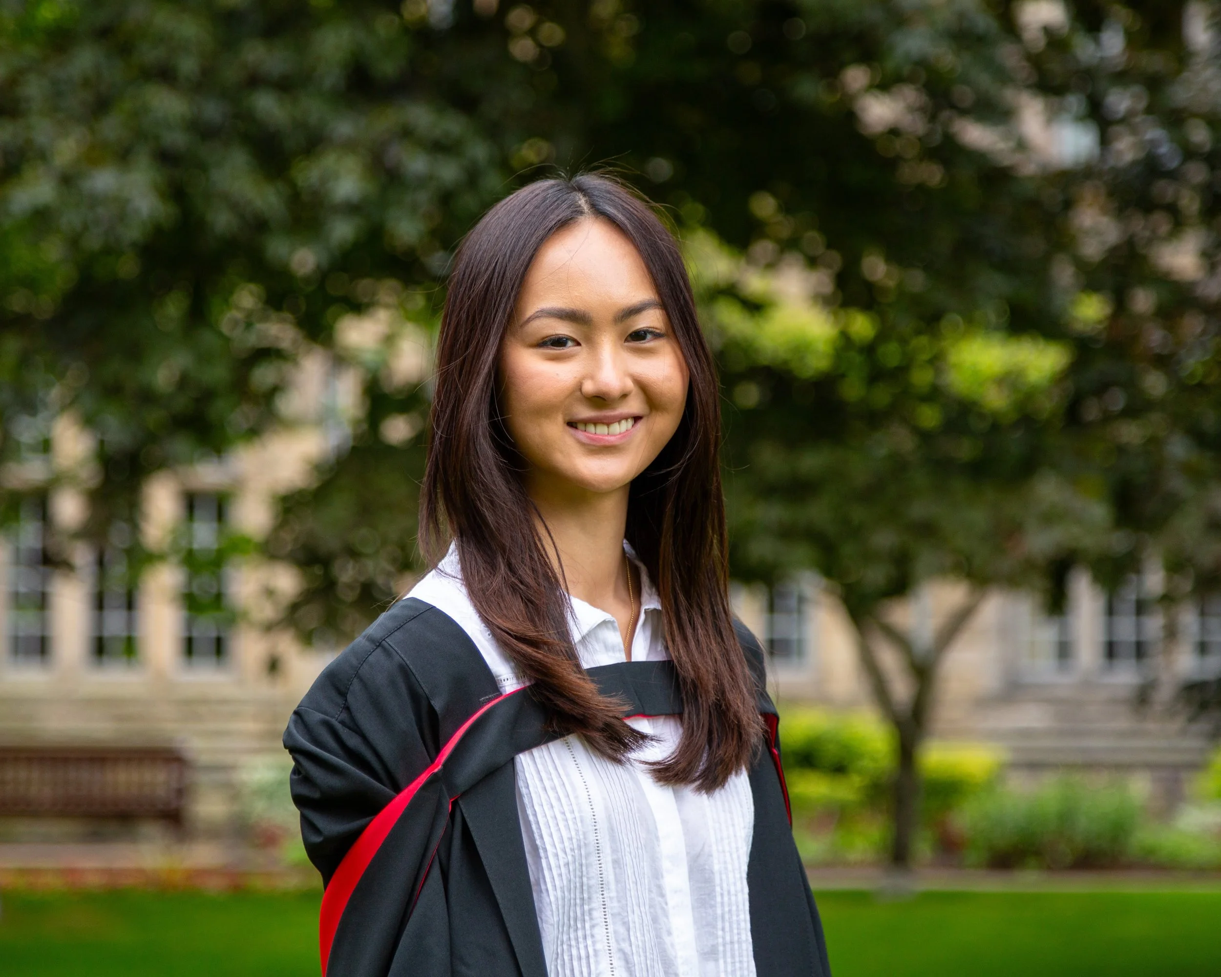 Young woman with long dark hair smiling outdoors, wearing a black graduation gown with red accents and a white button-up shirt, standing in front of green trees and a building.