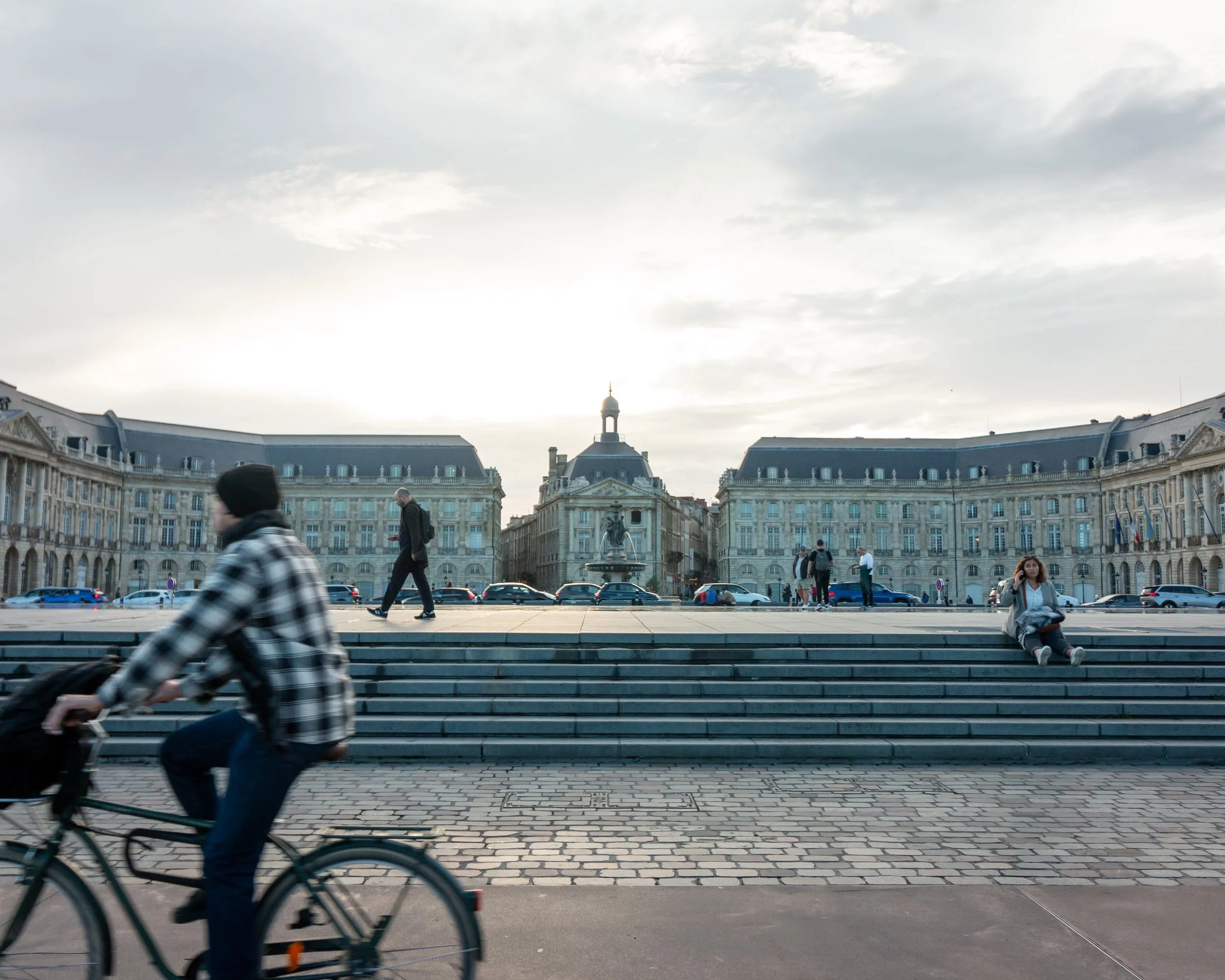 People walking and riding bikes in a city square with historic buildings and a fountain in the background during daytime.