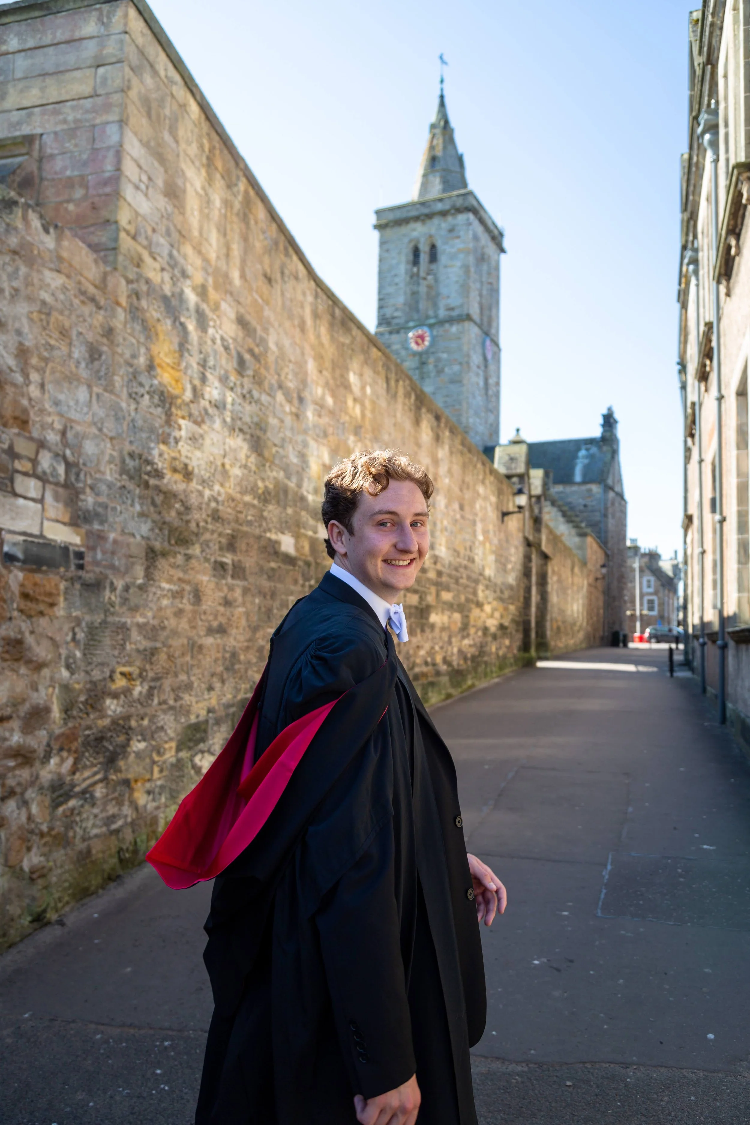 A young man in a black graduation gown and cap with a pink and black hood, smiling and walking outdoors on a sunny day with a historic stone church with a tall steeple in the background.